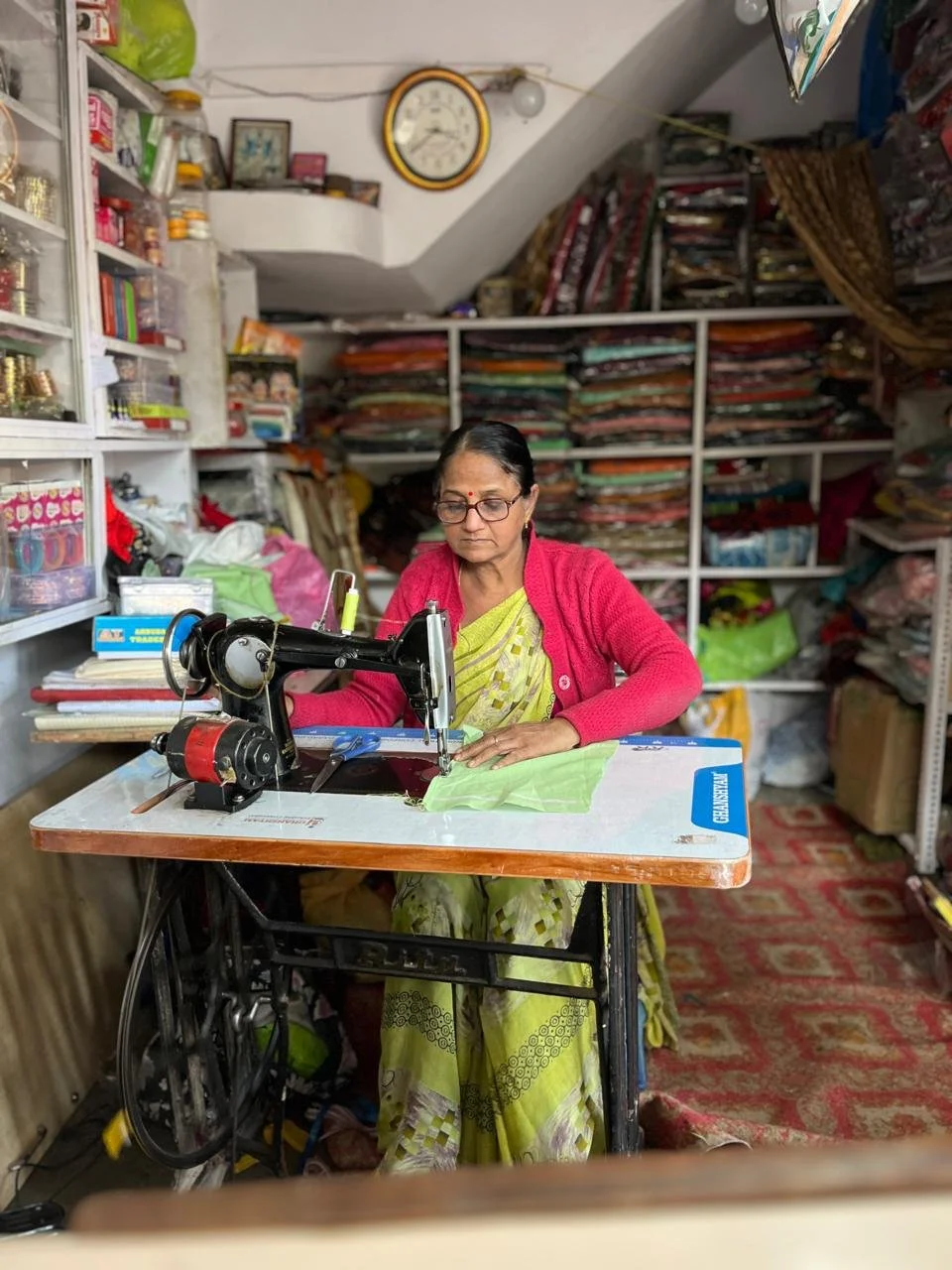 A woman sewing fabric on a sewing machine in a small shop with shelves filled with fabric and sewing supplies.