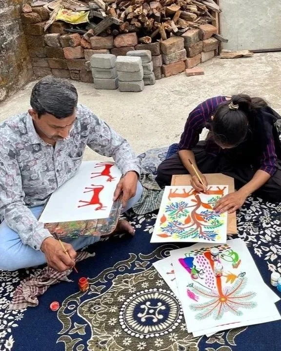 A man and a woman sitting on a black patterned rug outdoors, painting colorful designs on paper with markers and paints, with bricks and wood in the background.