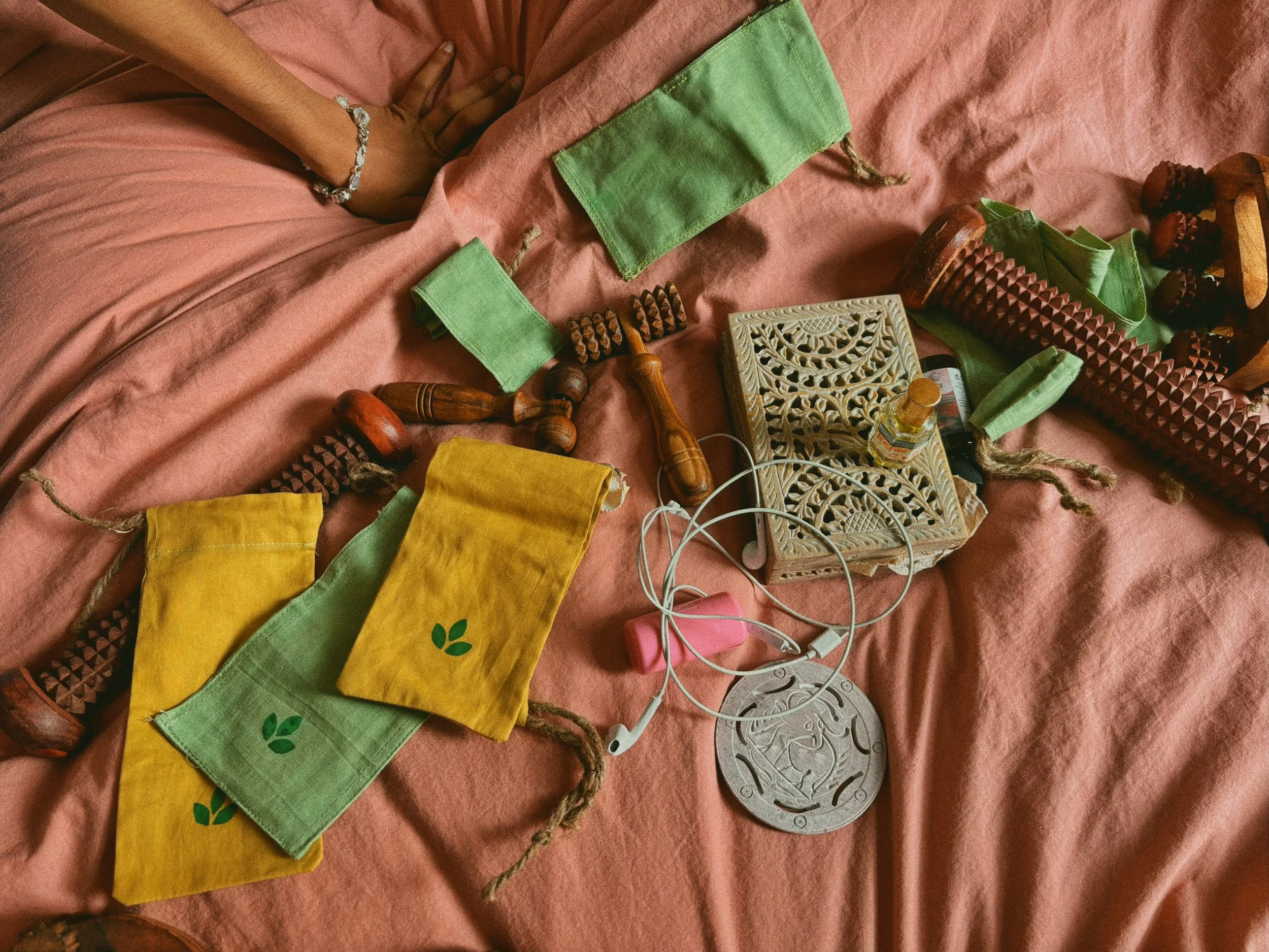 Wooden wellness massage tools are scattered aesthetically on a sheet. There are several cotton pouches with the Sarla logo screen printed on the fabric.