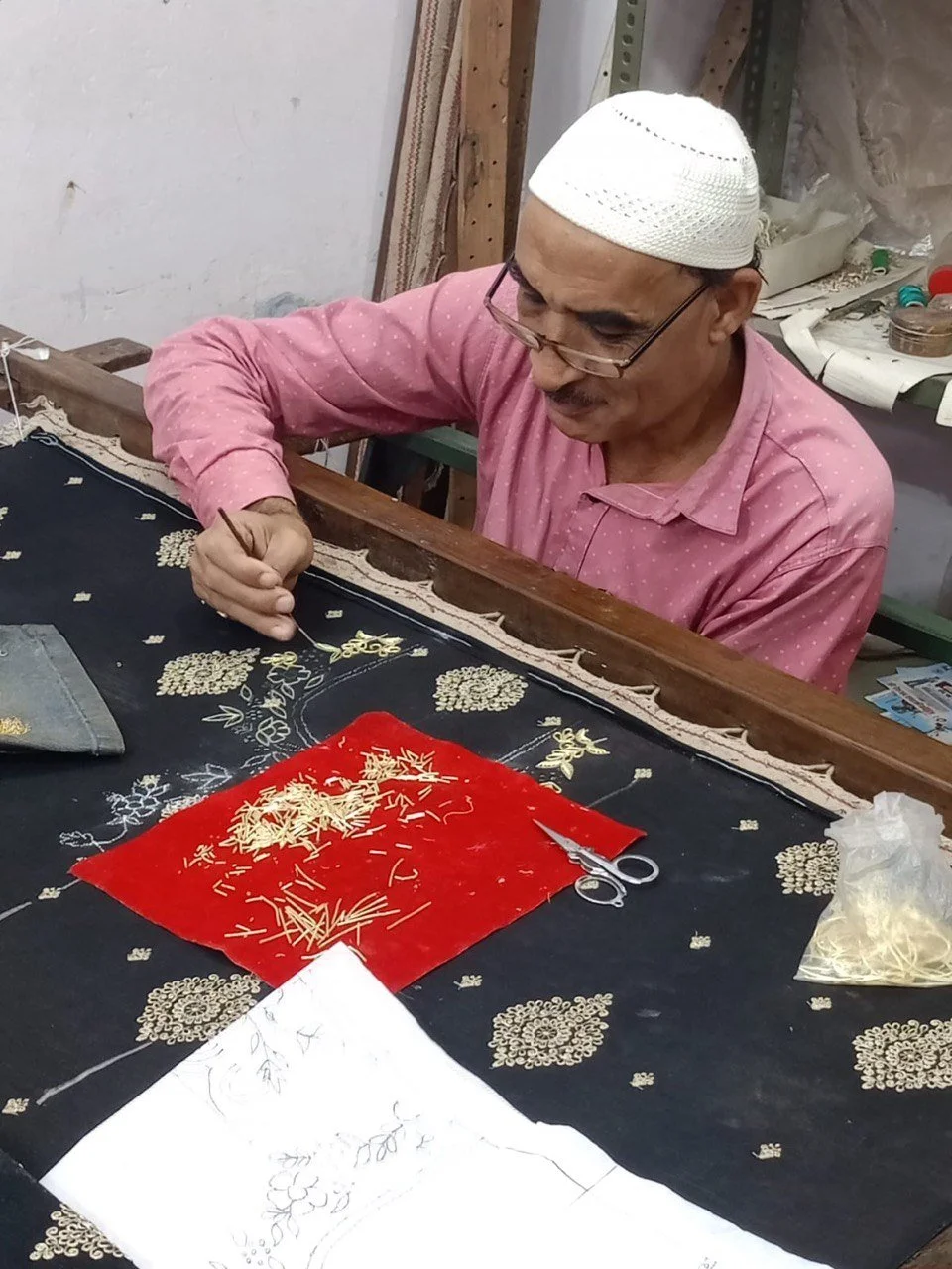 An elderly man wearing glasses and a white cap working on embroidery or stitching a fabric with a small needle, sitting at a worktable with various sewing supplies.