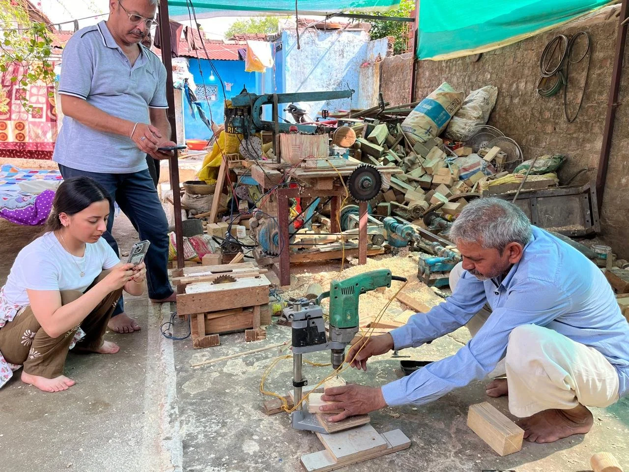 Four people working with woodworking tools outdoors, surrounded by wood scraps and machinery, one man is operating a drill while others are working in the background.