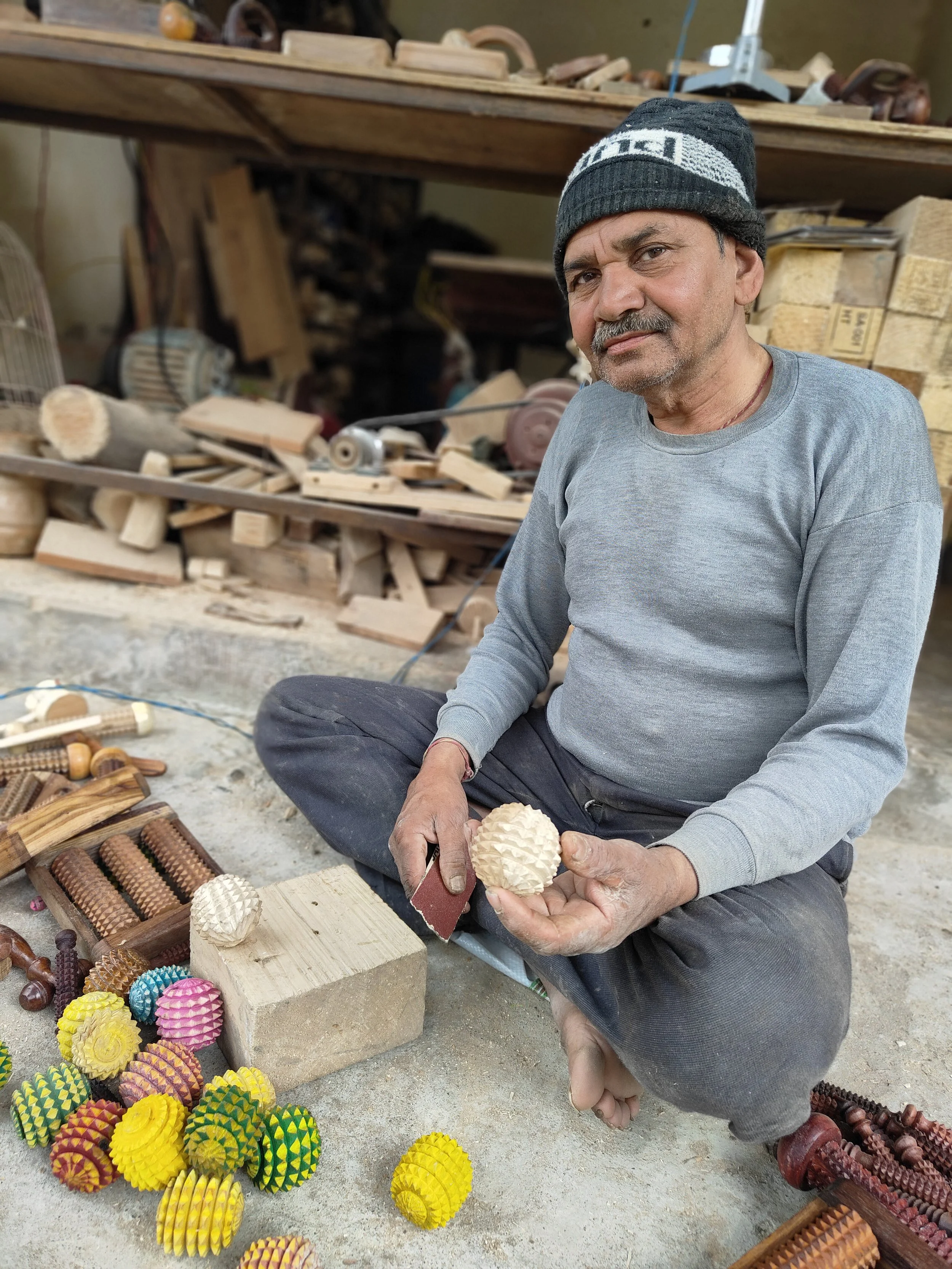 A man sitting on the ground in a workshop, holding a handmade wooden ball shaped object with spikes like a pine cone, surrounded by woodworking tools on shelves behind him.