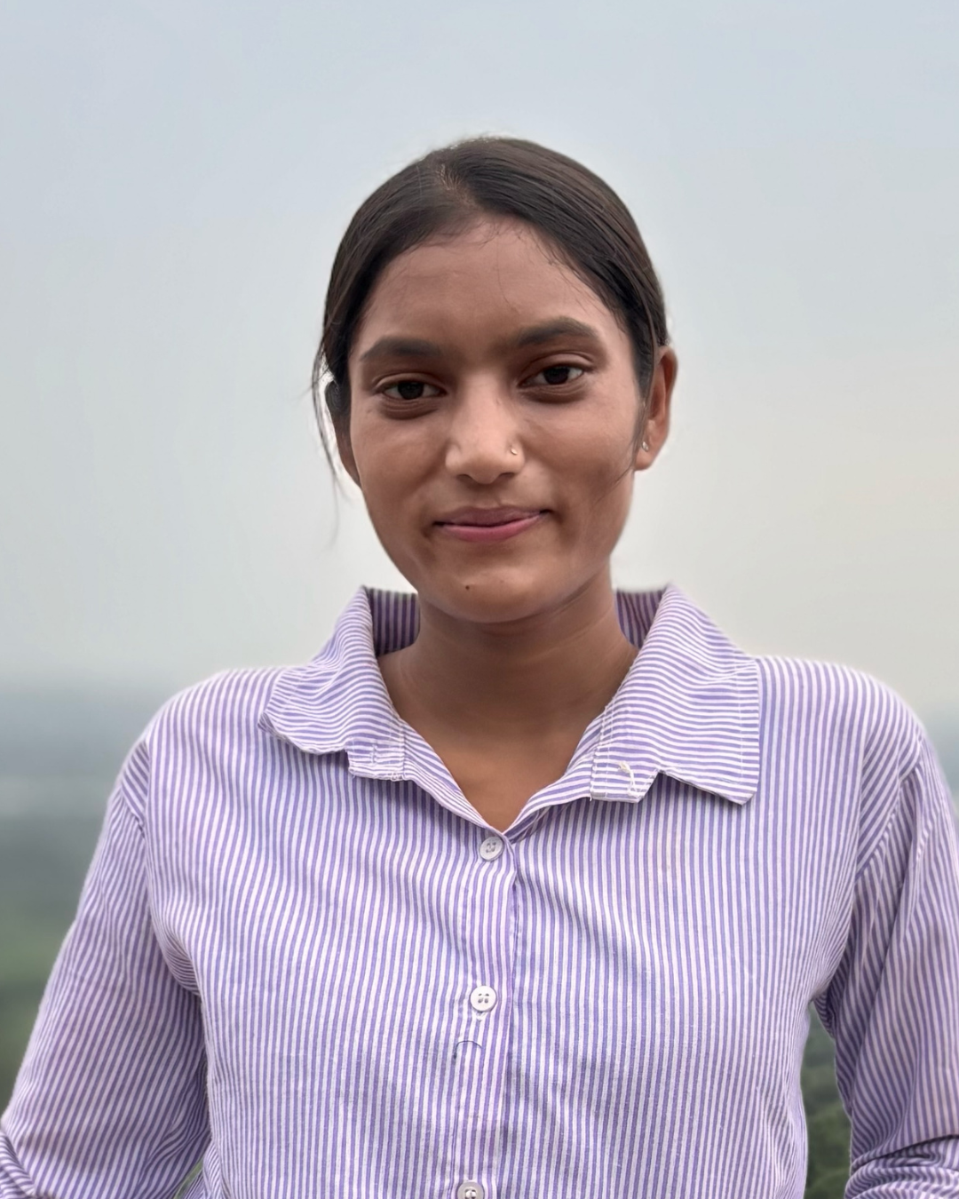 A girl wearing a blue and white striped shirt and looking into the camera and smiling. She is standing outside below clear blue skies.