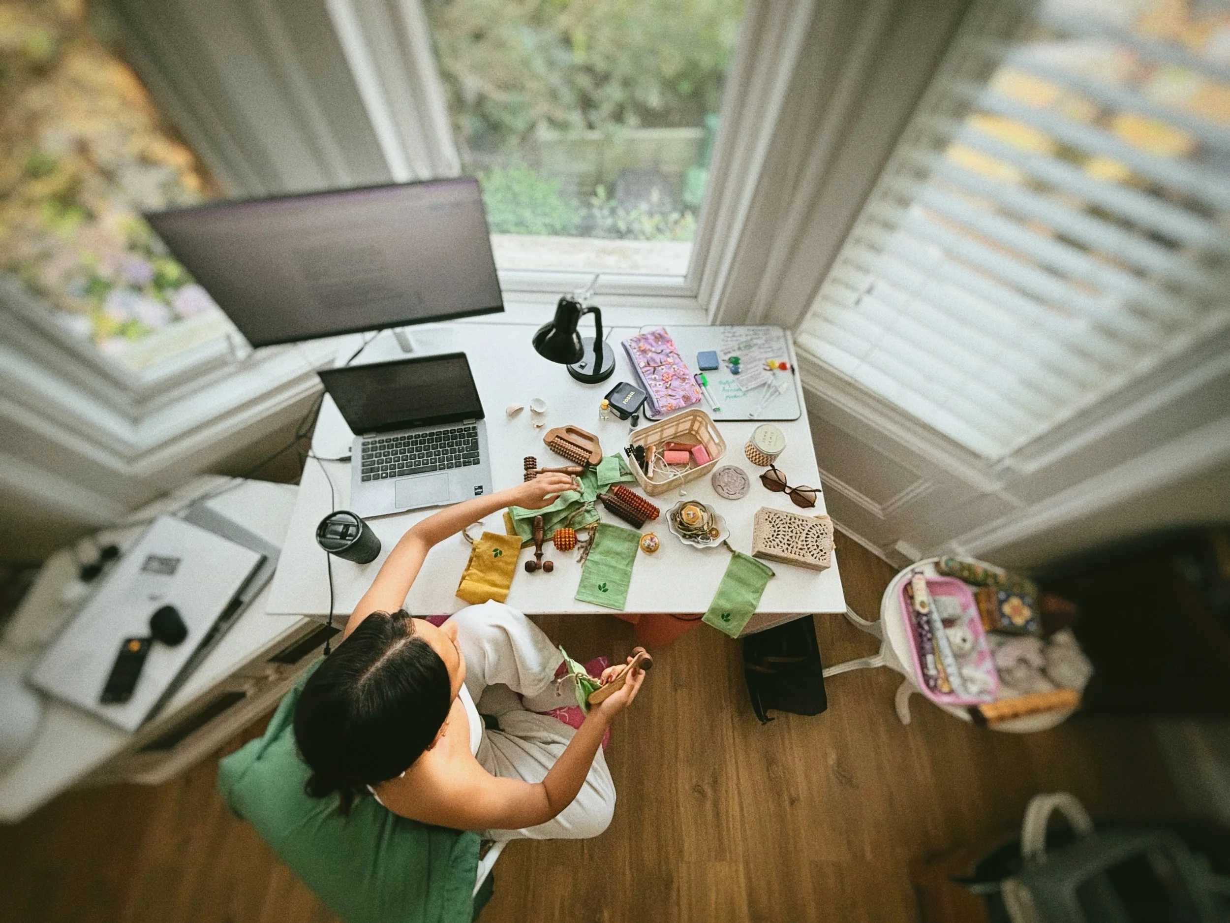 Founder working in her studio with all sarla products spread around the work station - from wooden wellness tools to packaging materials. There are three big windows and a huge desk with two monitor screens in front of her.