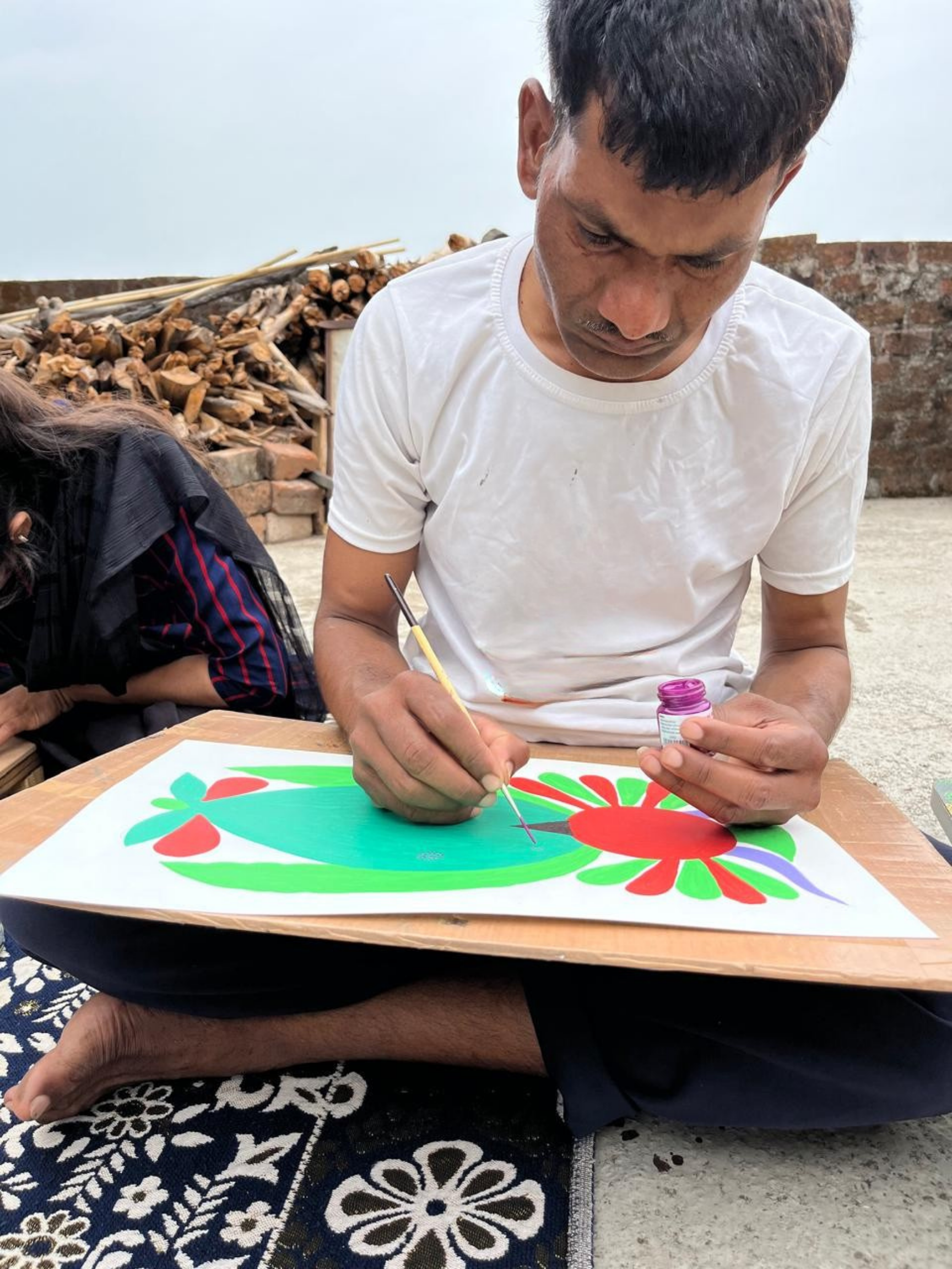Man painting a colorful design on paper with vibrant red, green, and purple colors, sitting outdoors.