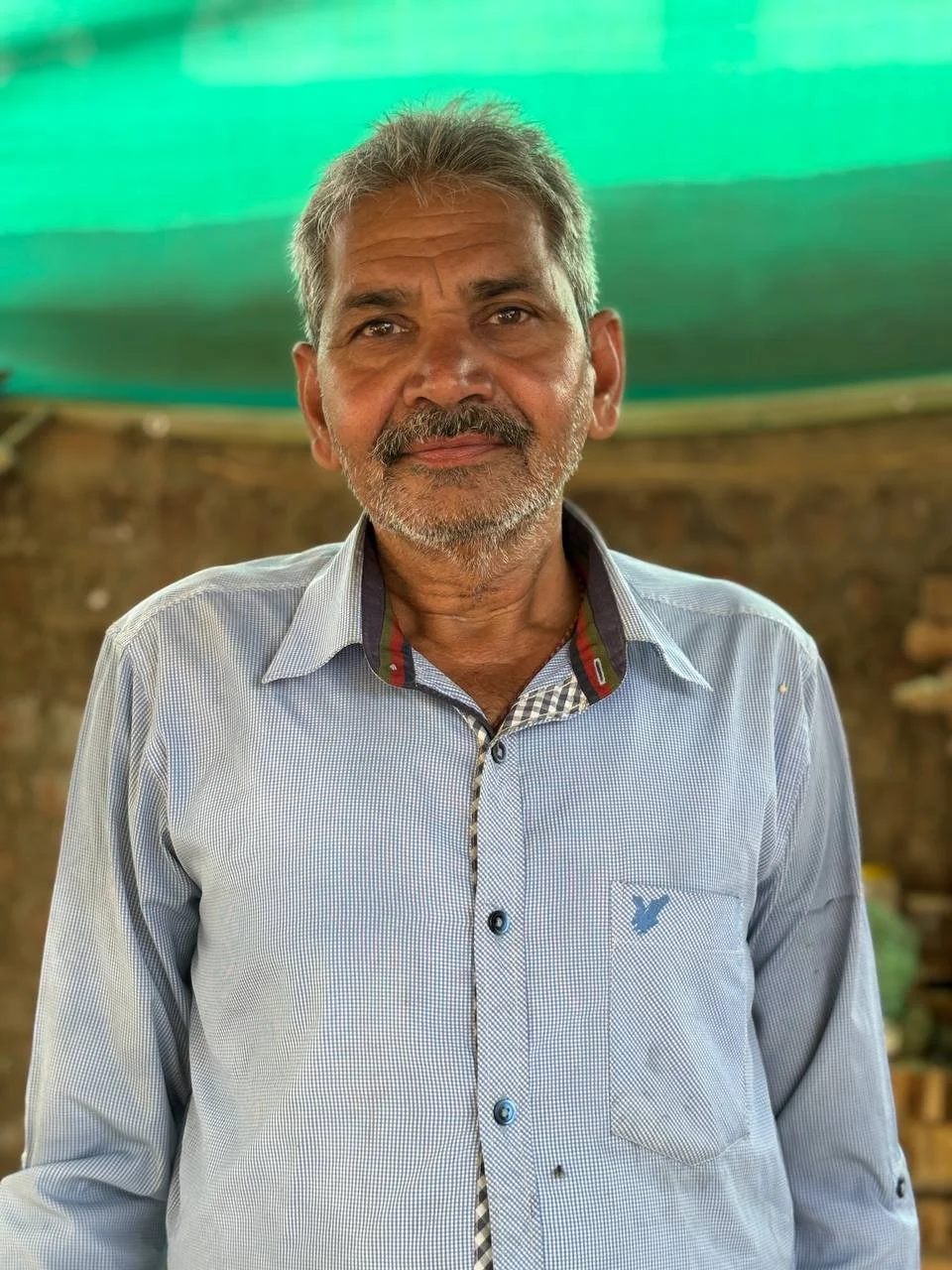 A middle-aged man with gray hair and a beard, wearing a light blue checkered shirt with an eagle logo on the chest, standing indoors with a green roof and brick wall background.