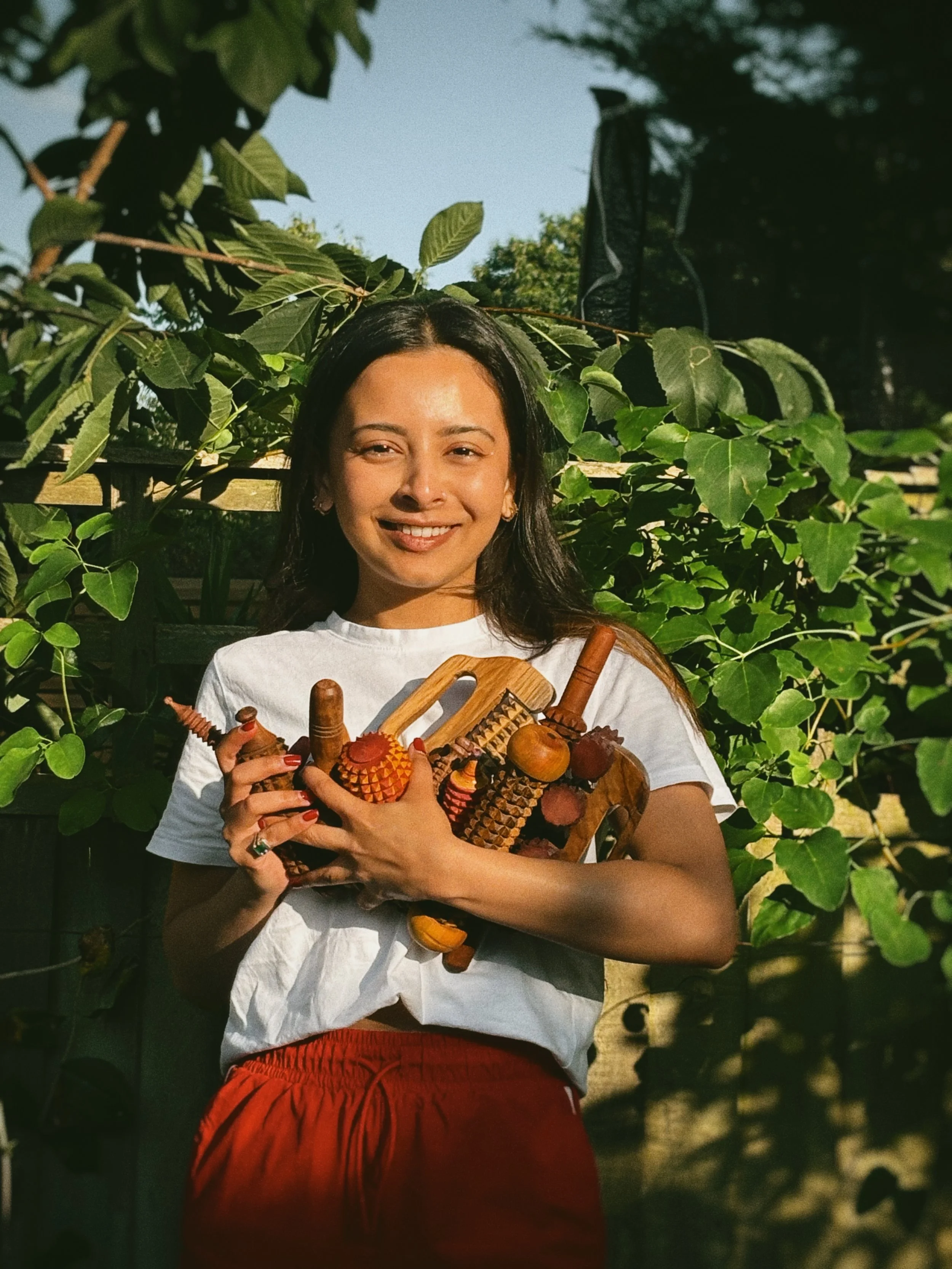 Founder holding all Sarla products in her arms close to her chest. She is wearing a white top and red pants and is standing in the middle of tall green trees.