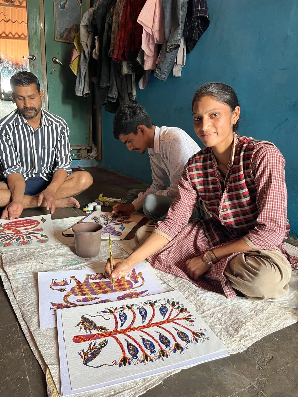 Three people sitting on the floor making traditional artwork on paper. One woman is painting a detailed, colorful design of a tree with animals. Two men are also working on similar artwork. Clothes hang on the wall behind them and a door is visible.