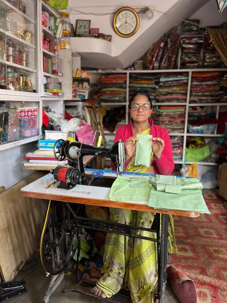 A woman sits at a sewing machine in a fabric shop, holding a piece of green cloth. She is wearing glasses and a red shawl with traditional Indian attire. The shop has shelves filled with colorful fabrics and sewing supplies.