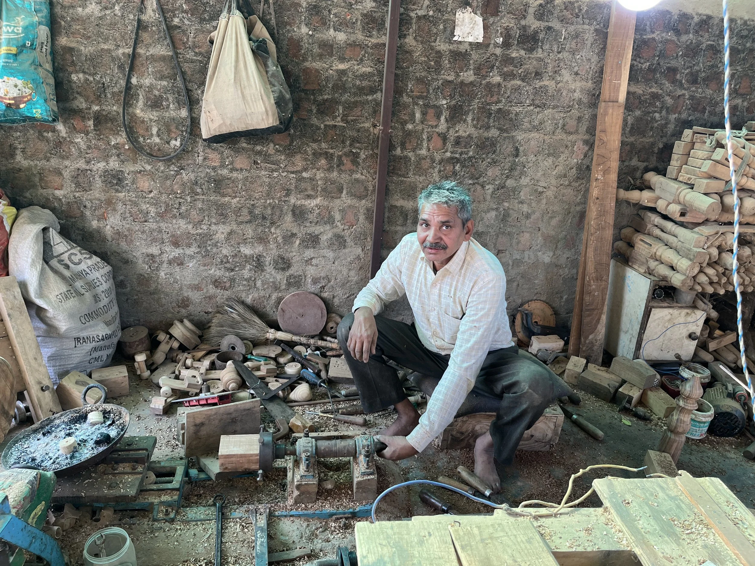 A man with gray hair and a mustache, wearing a light-colored checkered shirt and black pants, crouching down in a woodworking shop surrounded by wood shavings, tools, and wooden pieces. The background features a brick wall with hanging bags and a stack of rolled or carved wooden sticks.