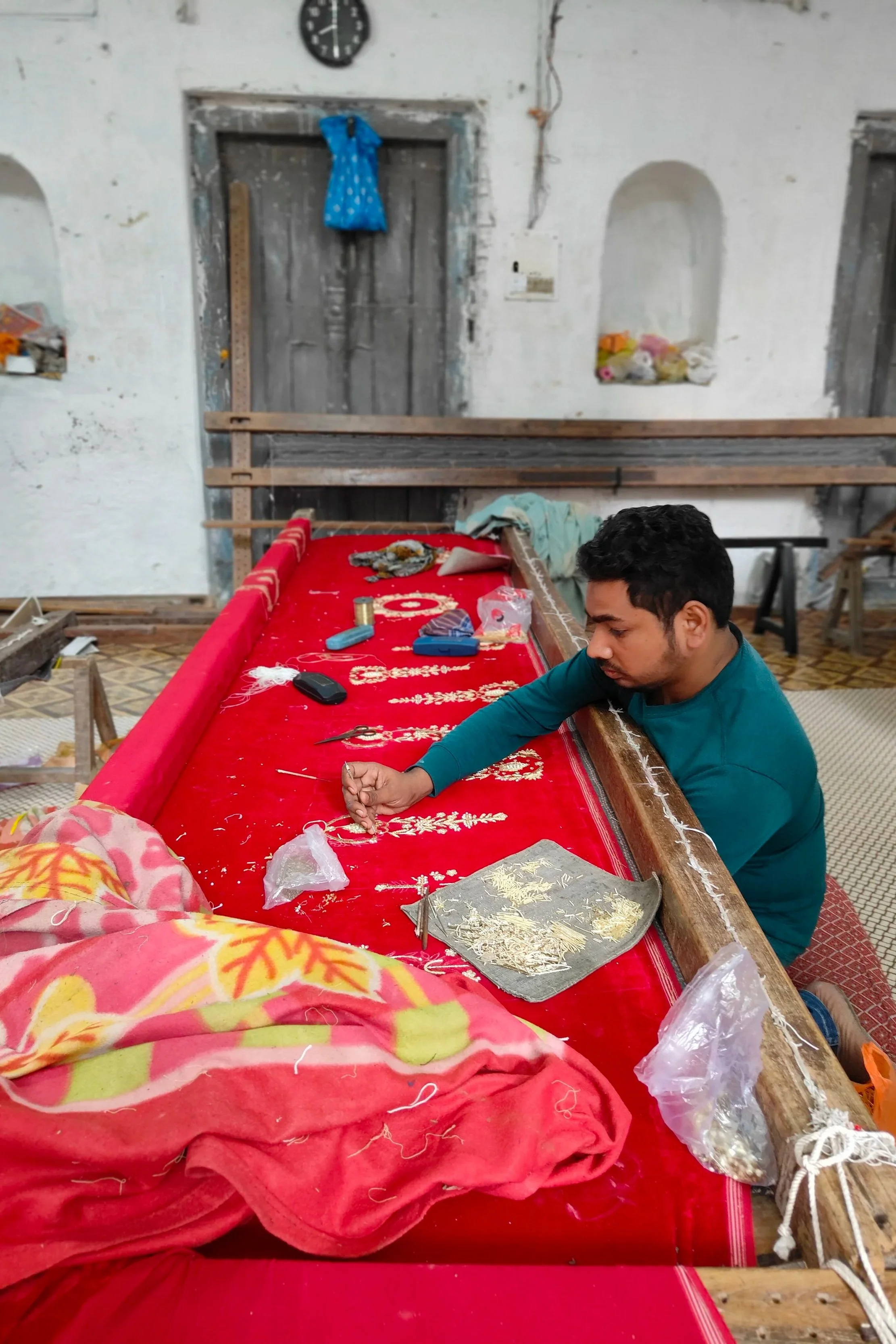 A man sitting on the floor, working on embroidery or craft on a red cloth laid on a low wooden frame, in a rustic room with white walls, a wall clock, and various tools and materials scattered around.