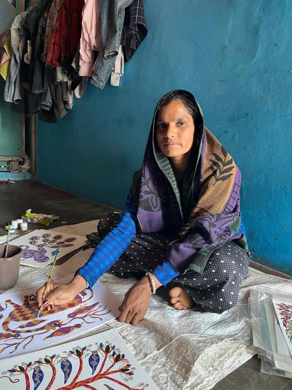A woman sitting on a floor, painting traditional artwork on a sheet of paper. She is wearing a colorful shawl and polka-dot pants, with a blue checkered shirt underneath. There are various painted artworks on the ground in front of her and a blue wall behind her with hanging clothes.