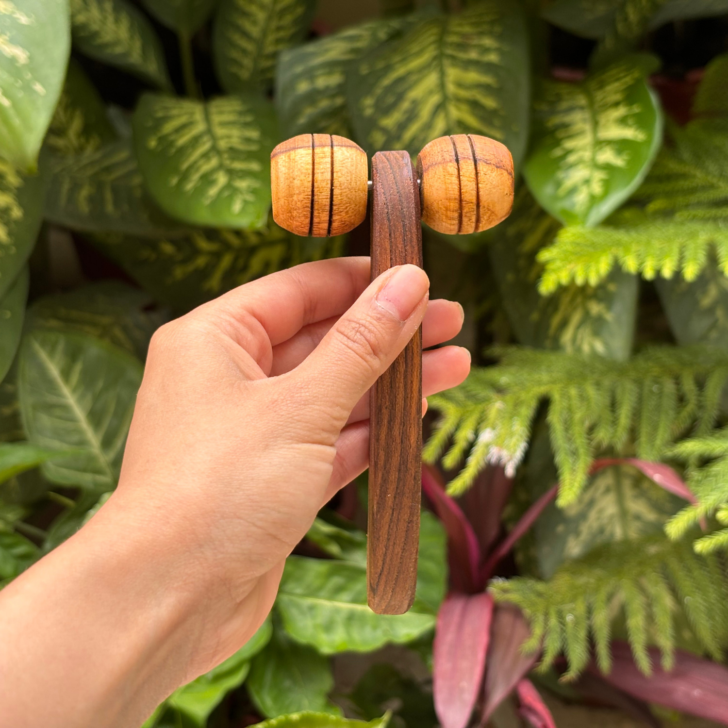 Wooden lymphatic face roller held in hand against a natural leafy background, showing its compact handheld design.