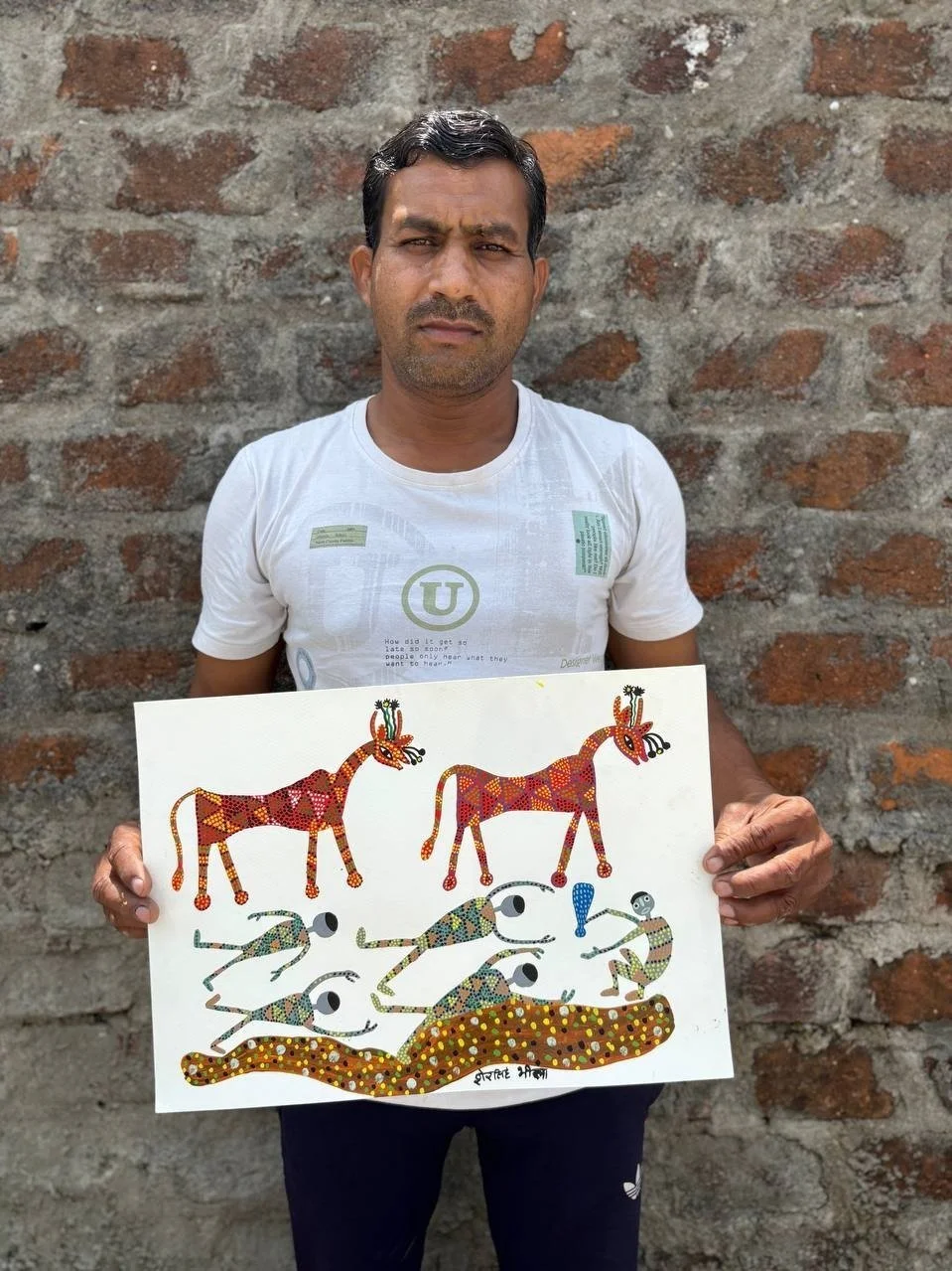 Man standing in front of a brick wall, holding a colorful drawing of stylized animals, including giraffes and other creatures.