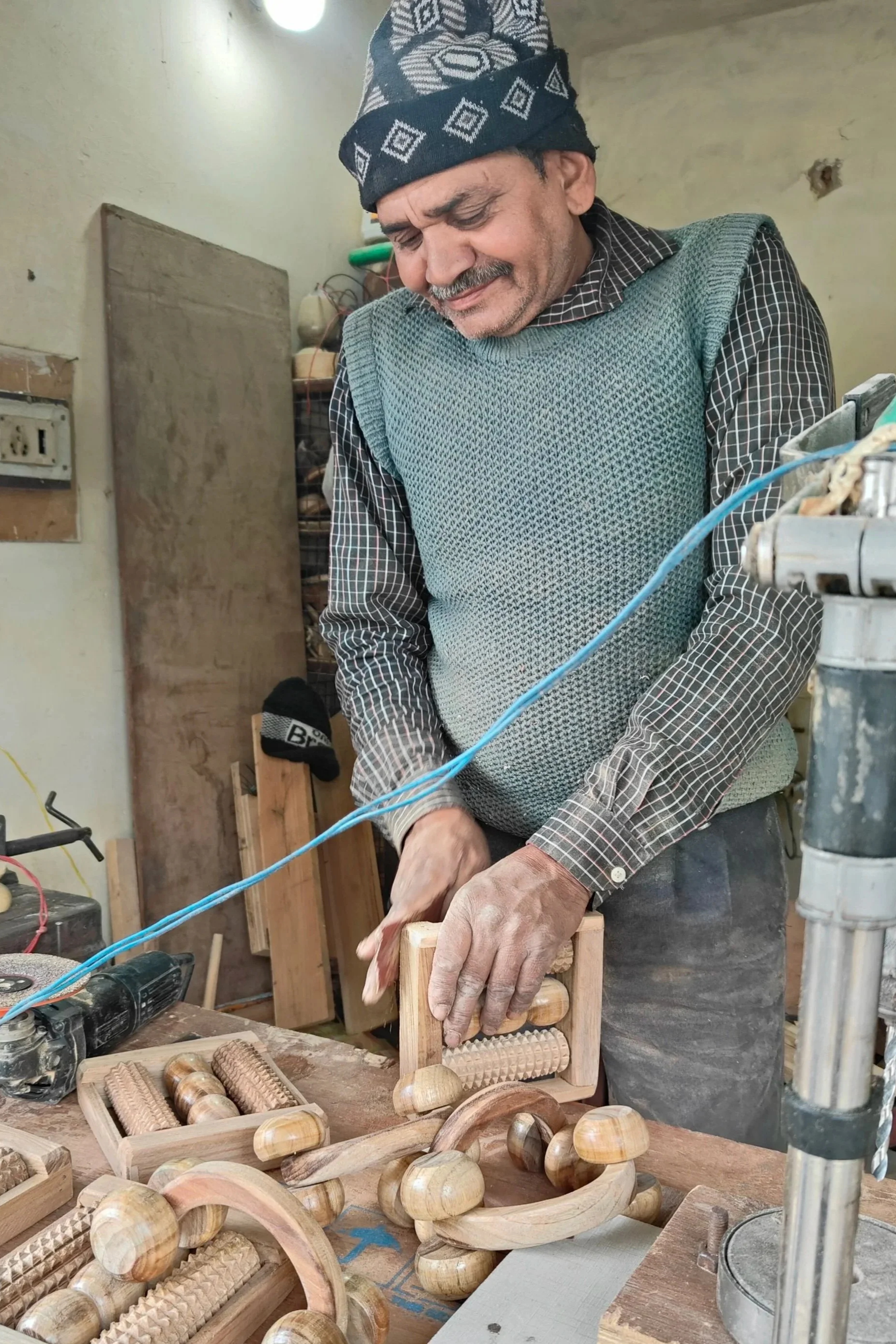 A man working on a woodworking project in a workshop, surrounded by wooden toys and tools, wearing a patterned headscarf and a checkered shirt.