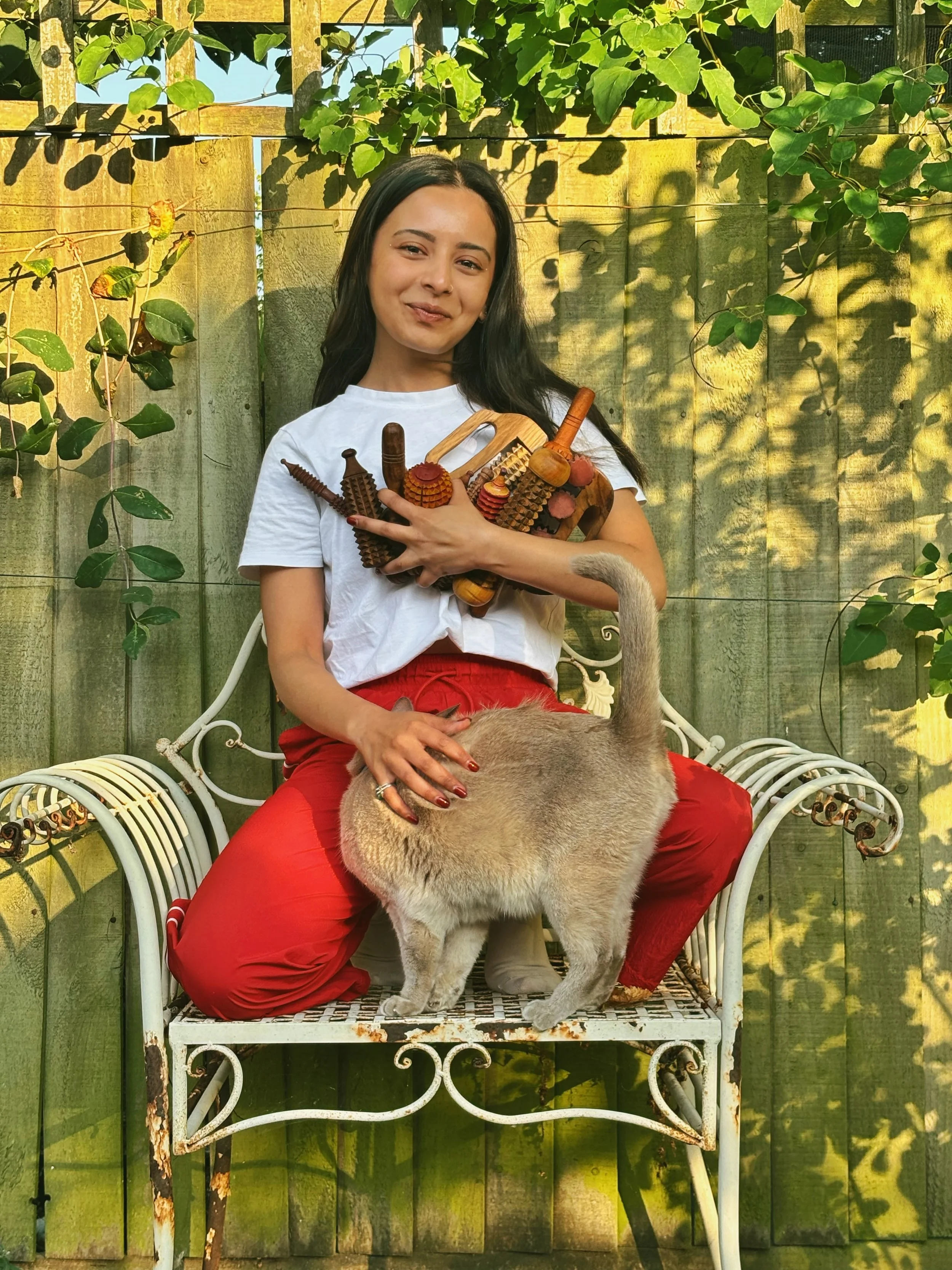 A young woman holding all wooden products in her arms close to her chest looking into the camera and smiling. She is wearing a white top and red pants and is surrounded by green trees.