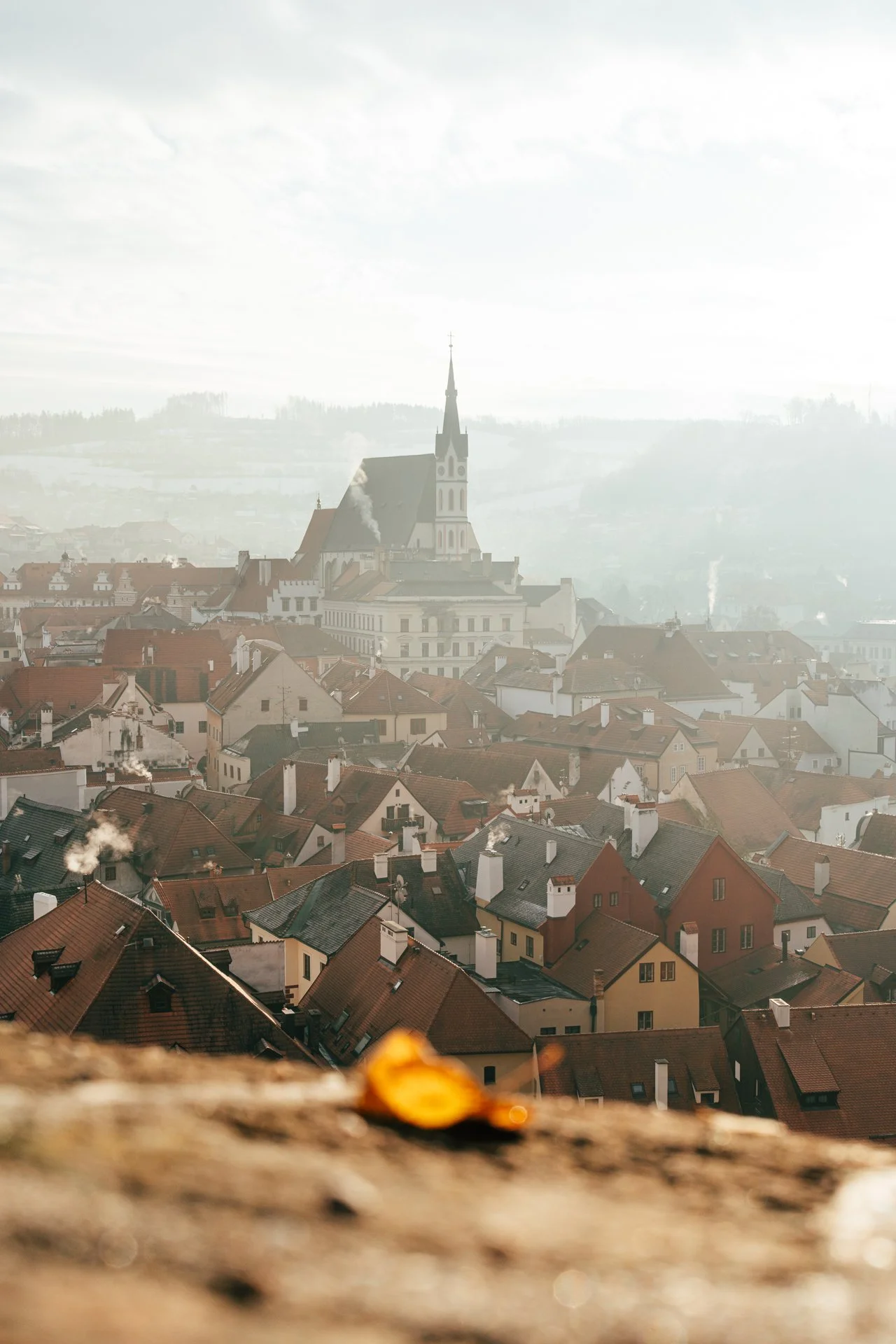 Aerial view of a European town with a prominent church tower, surrounded by red-roofed buildings and a misty landscape.