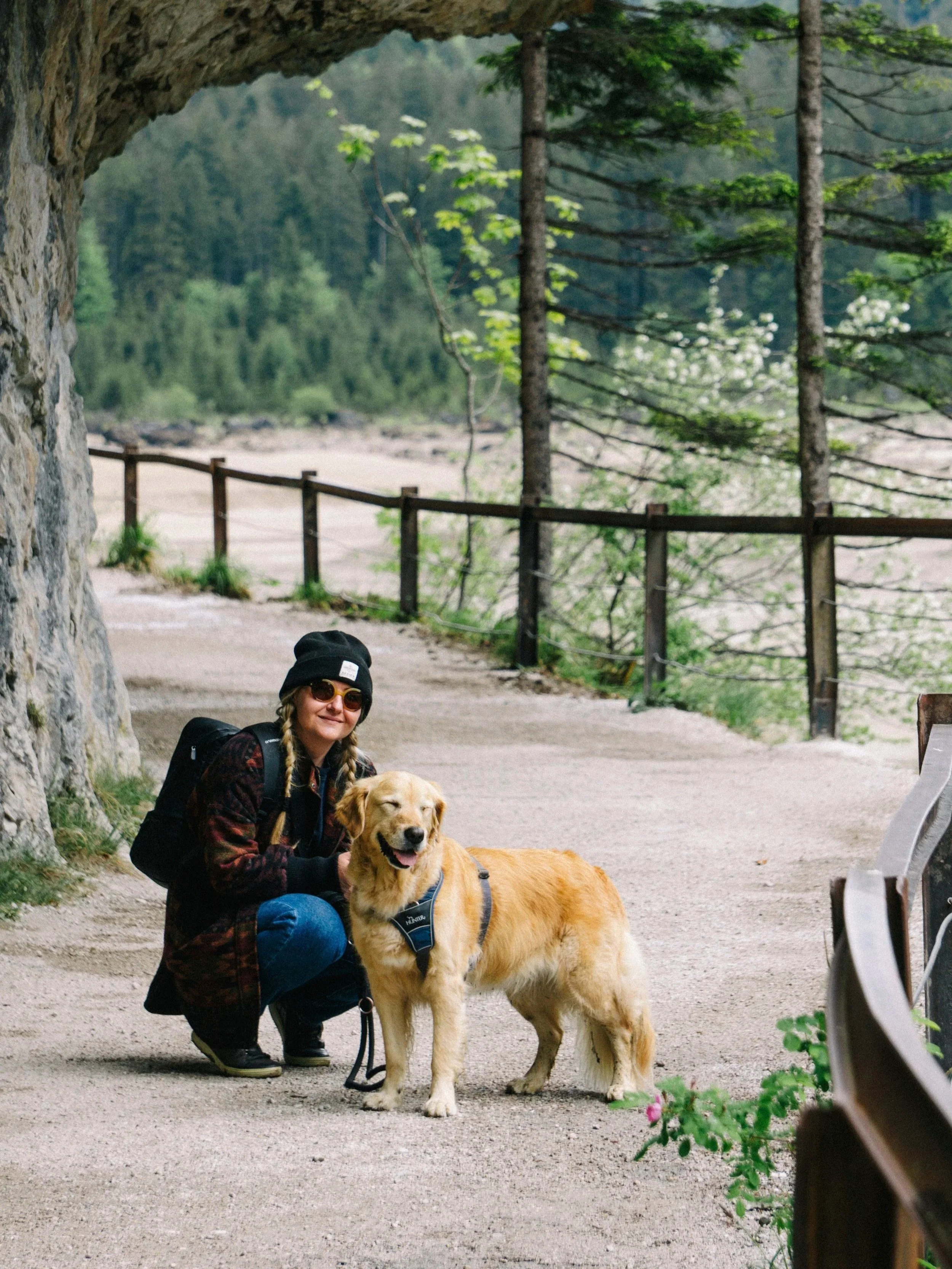 A woman wearing sunglasses, a black beanie, and a checkered jacket squats down next to a golden retriever dog on a leash. The woman is smiling, and the dog appears happy with its tongue out. They are outside under a rocky archway with tall trees and a wooden fence in the background.