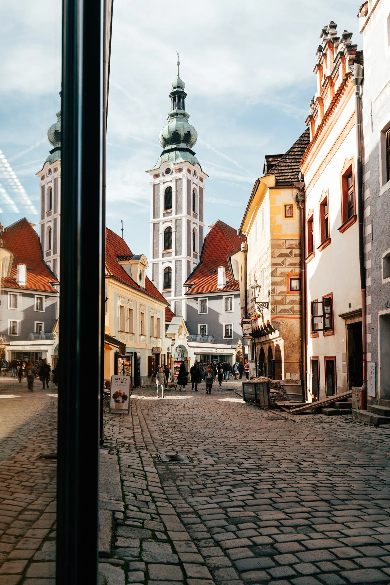 Historic European street with cobblestones, old buildings, and a church with a tall spire, featuring people walking and a gelato sign.