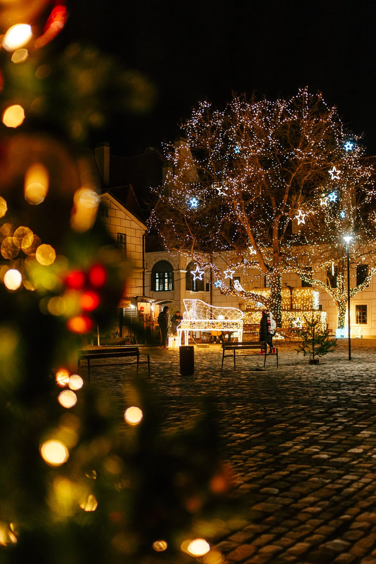 Christmas decorations in a city square at night with illuminated trees and a large light structure shaped like a piano. Blurred foreground with festive lights, people walking near benches, and historic buildings in the background.