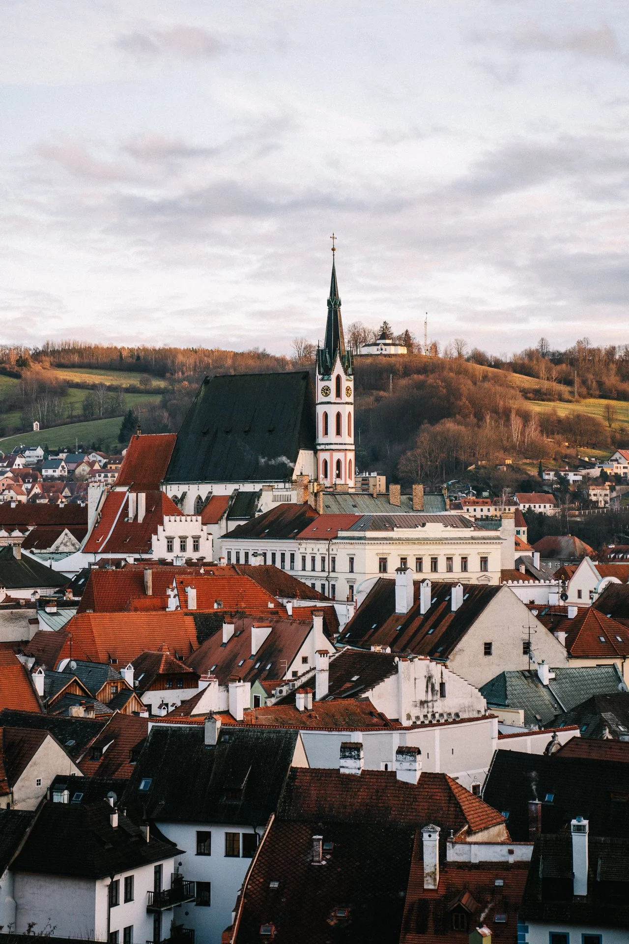 Scenic view of a historic European town with a church steeple and red-roofed buildings, set against a backdrop of rolling hills and a cloudy sky.