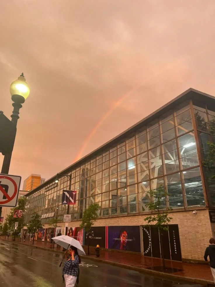 A city street scene with a woman holding an umbrella, wet pavement, and a glass building with illuminated interior lights. The sky is pinkish with a faint rainbow visible.