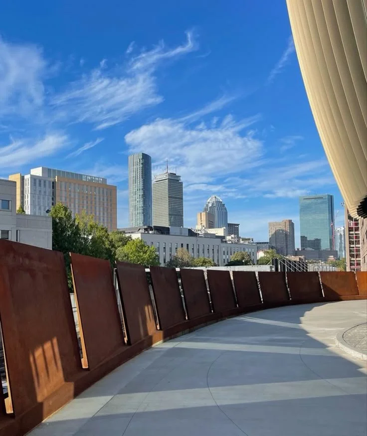 View of a city skyline with tall buildings under a bright blue sky with wispy clouds, viewed from Northeastern EXP Bridge, with the signature curved rust-colored railing and a large beige curtain partially visible on the right side.