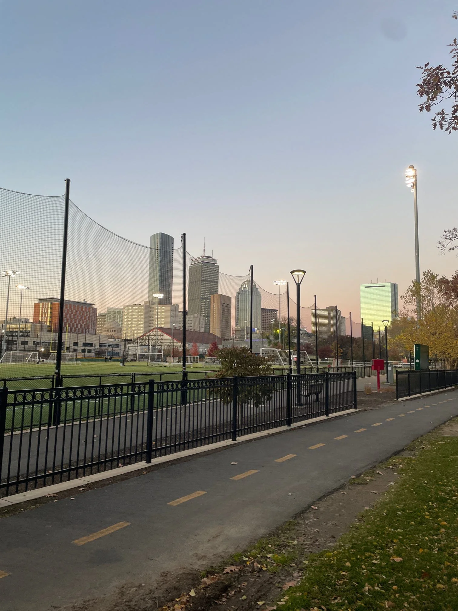 A city skyline viewed from a park with a walking path, a fenced sports field, and tall floodlights, with a few trees and a clear sky at dusk.
