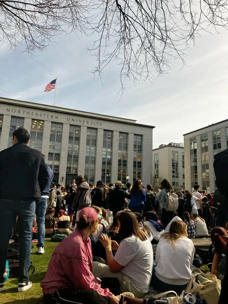 Crowd gathered outside Northeastern University campus building on a sunny day with leafless tree branches above.