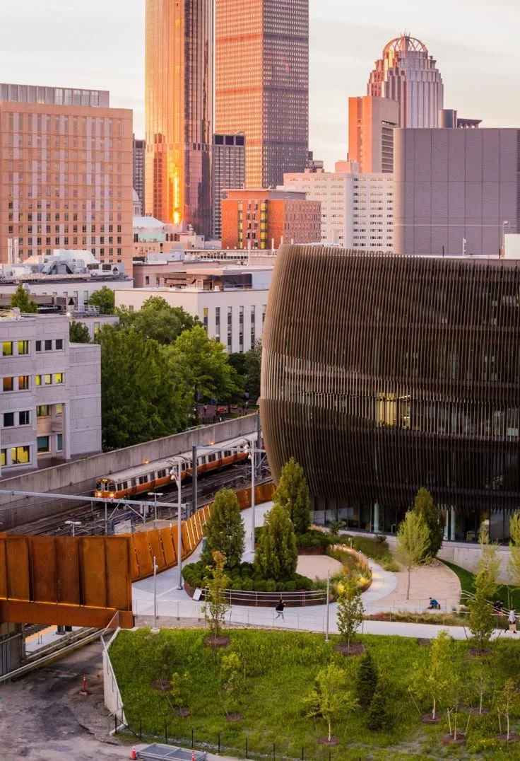 Sunset cityscape view of Northeastern EXP and ISEC from across the orange line train track, with modern skyscrapers, a train on elevated tracks, and a curved black building surrounded by green trees and a park.