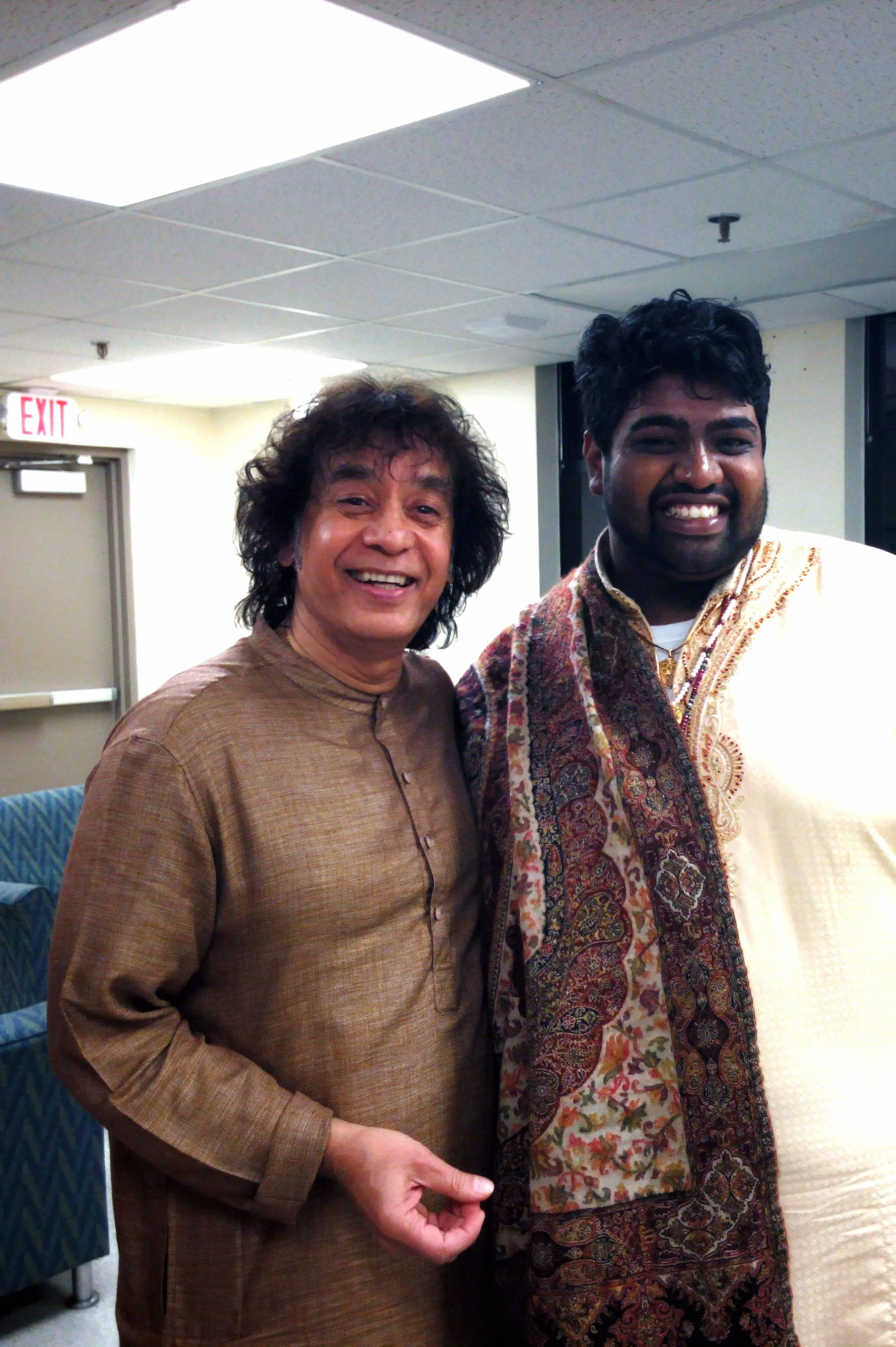 Two people smiling, standing together indoors, wearing traditional attire. The person on the left is in a brown kurta, and the person on the right is in an embroidered cream outfit with a colorful shawl. A door and a couch are in the background.