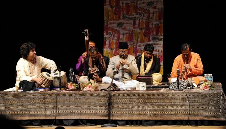 Musicians performing traditional Indian music on stage, with instruments like tabla and sitar. The setting is decorated with a patterned cloth backdrop, and microphones are placed in front.