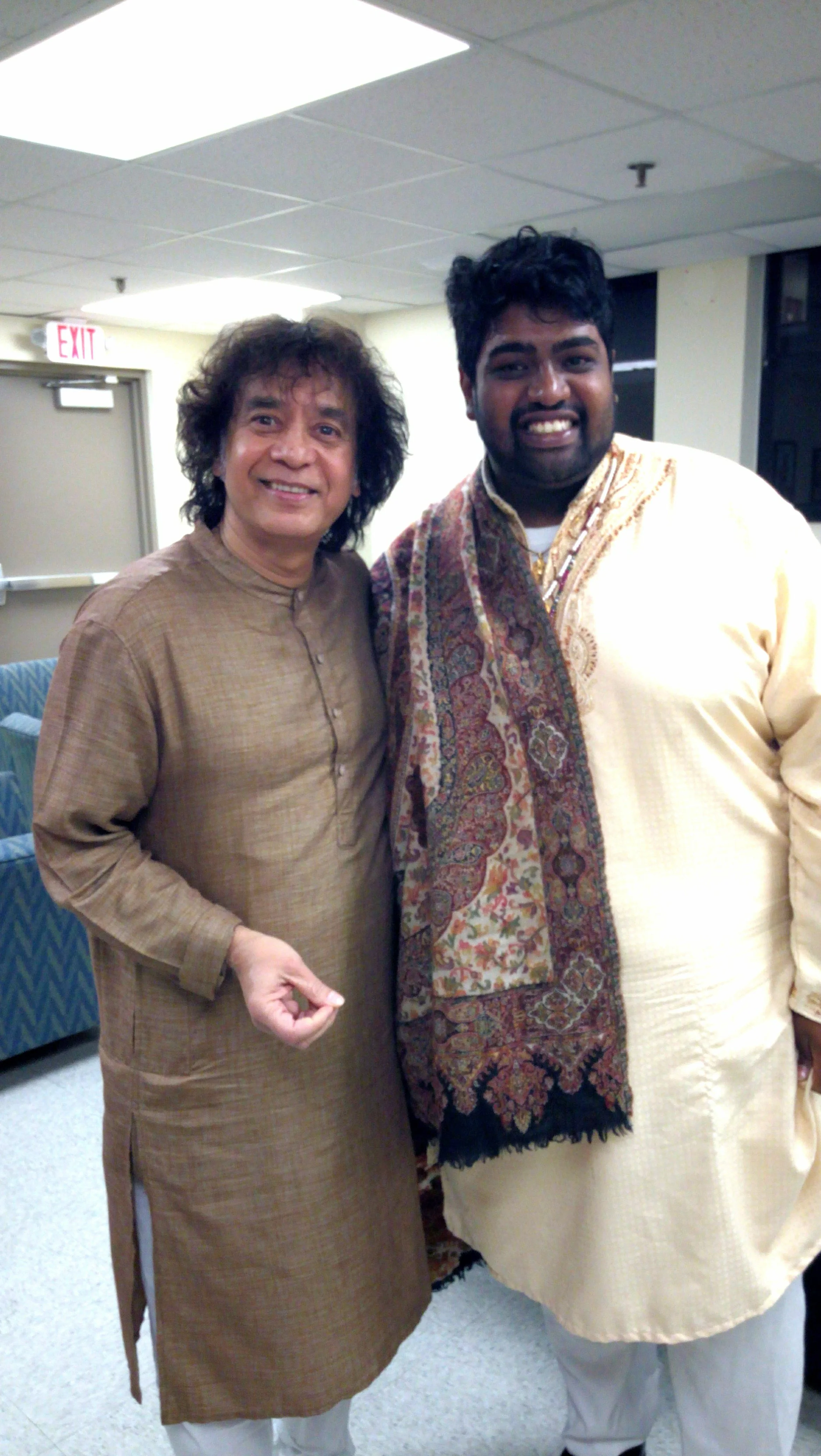 Two men in traditional attire standing together indoors, one wearing a brown kurta and the other wearing a beige outfit with a patterned shawl.