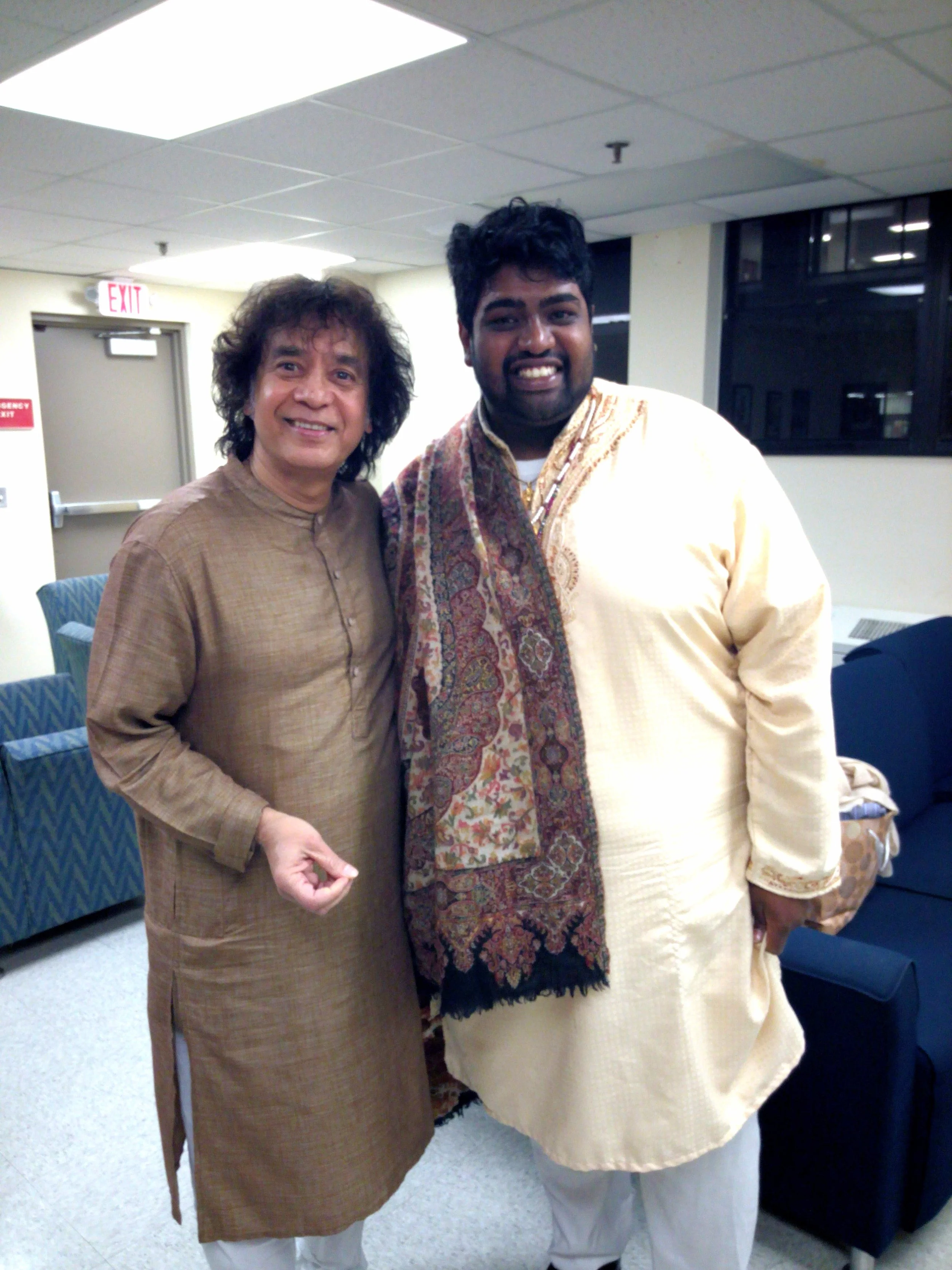 Two men in traditional Indian attire standing together in a room with a couch and overhead lighting.