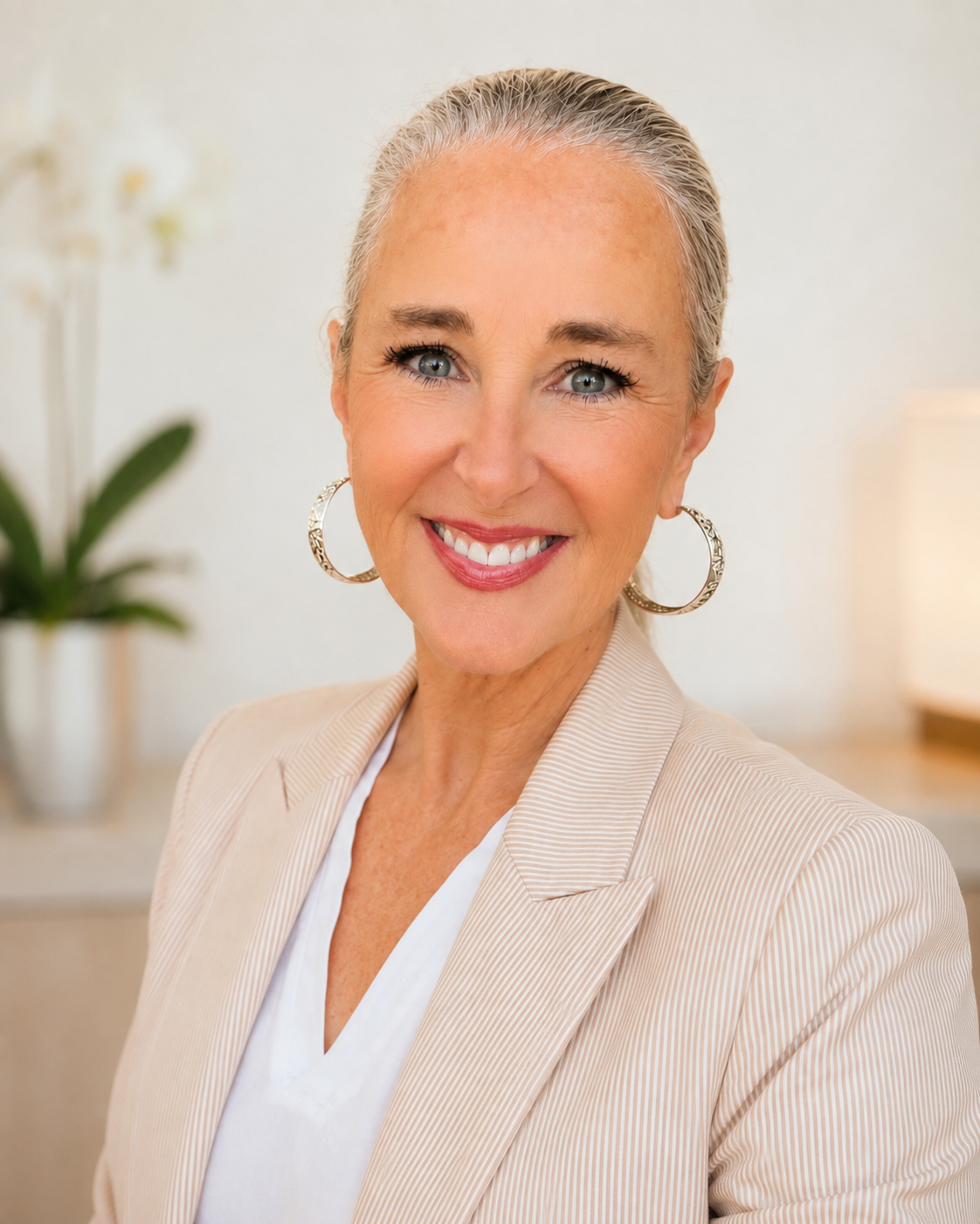 A portrait of a smiling mature woman with gray hair pulled back, wearing hoop earrings and a light-colored blazer, in a well-lit indoor space with a blurred background including a white orchid plant.
