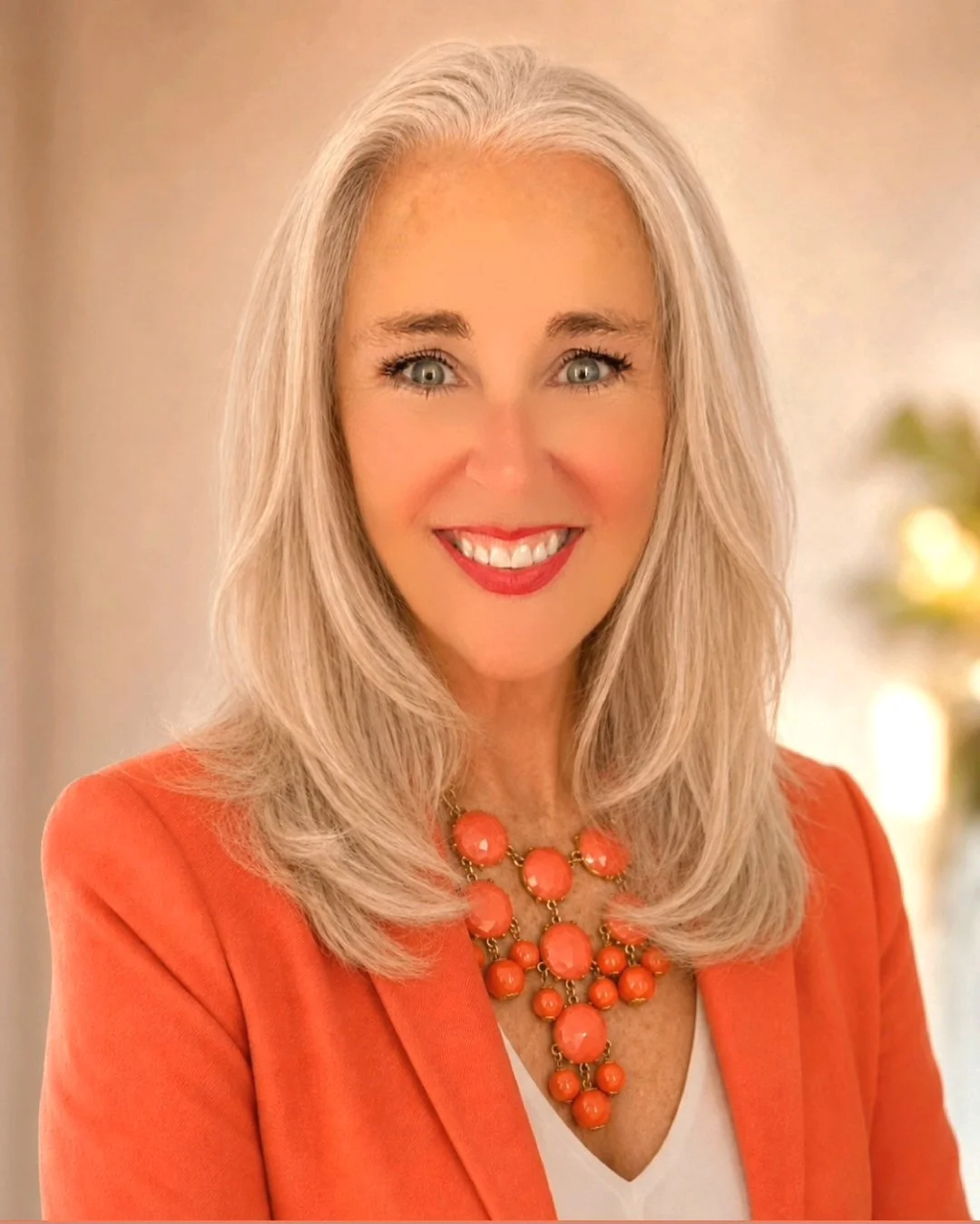 A smiling woman with blonde hair wearing an orange blazer and matching statement necklace.