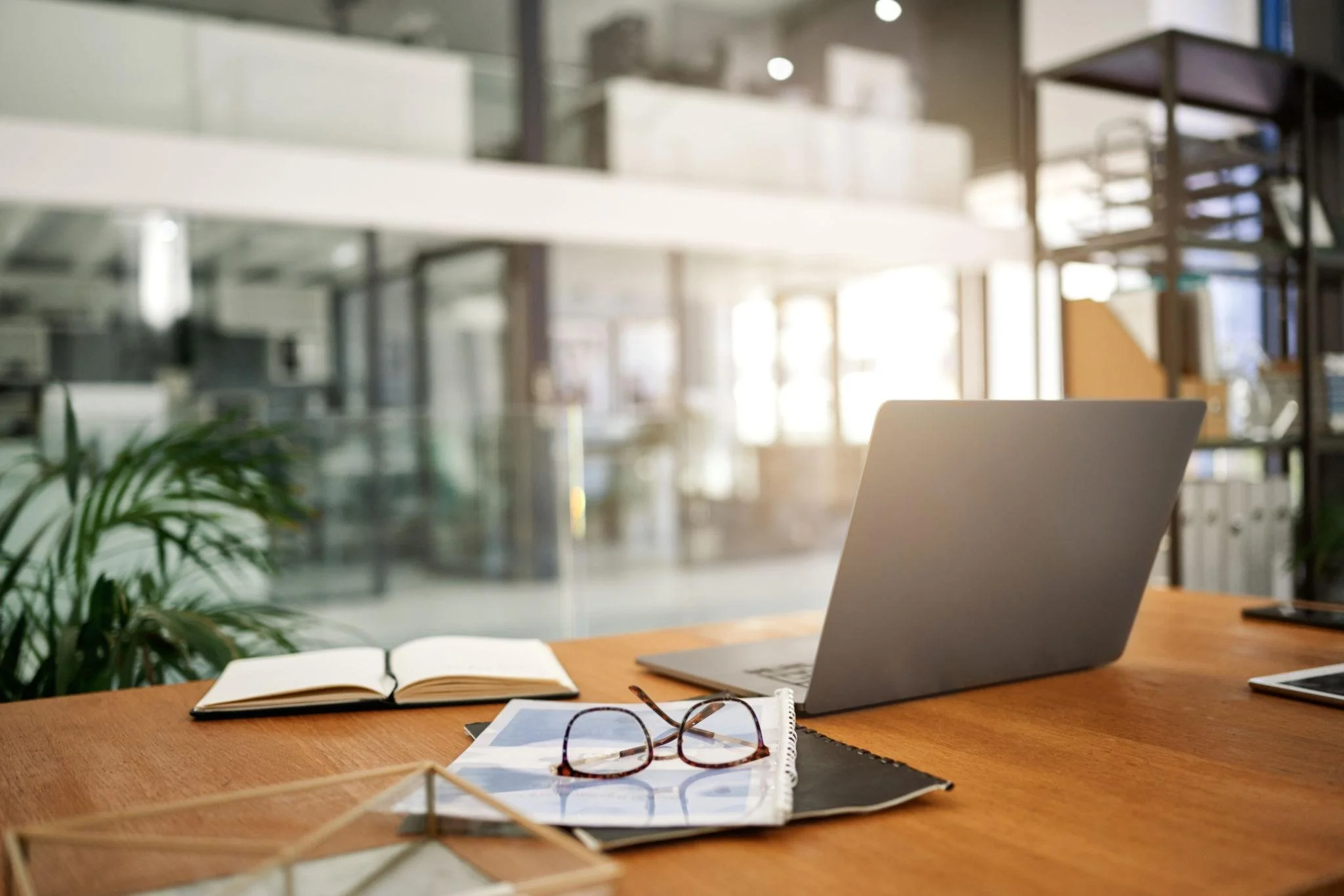 A workspace with a laptop, eyeglasses, an open notebook, and printed documents on a wooden desk, with a blurred background of an office or library setting.