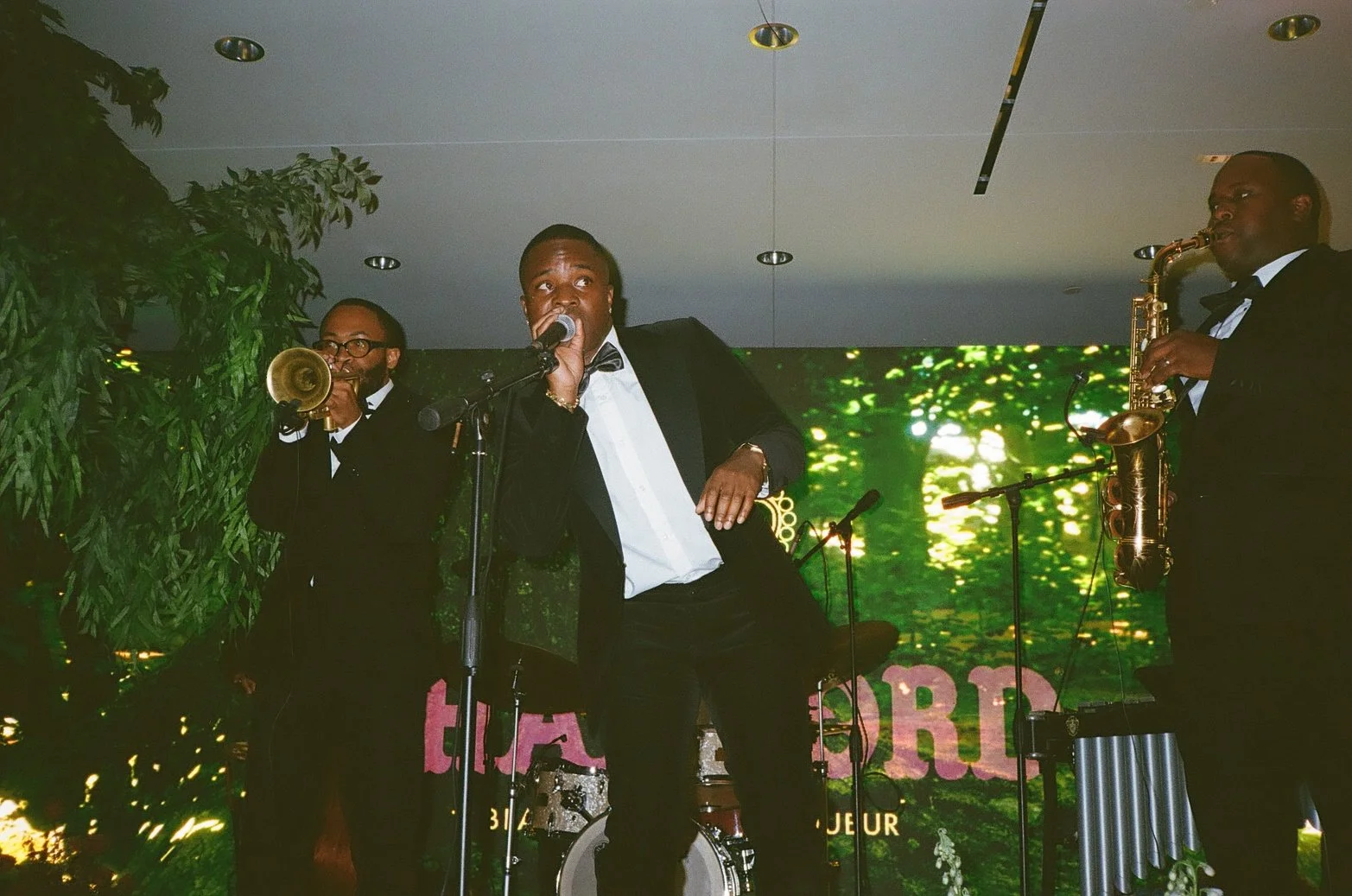 Three men in tuxedos performing on stage, with one singing into a microphone, a man playing trumpet, and another playing saxophone. They are under stage lighting with a green nature-themed background.