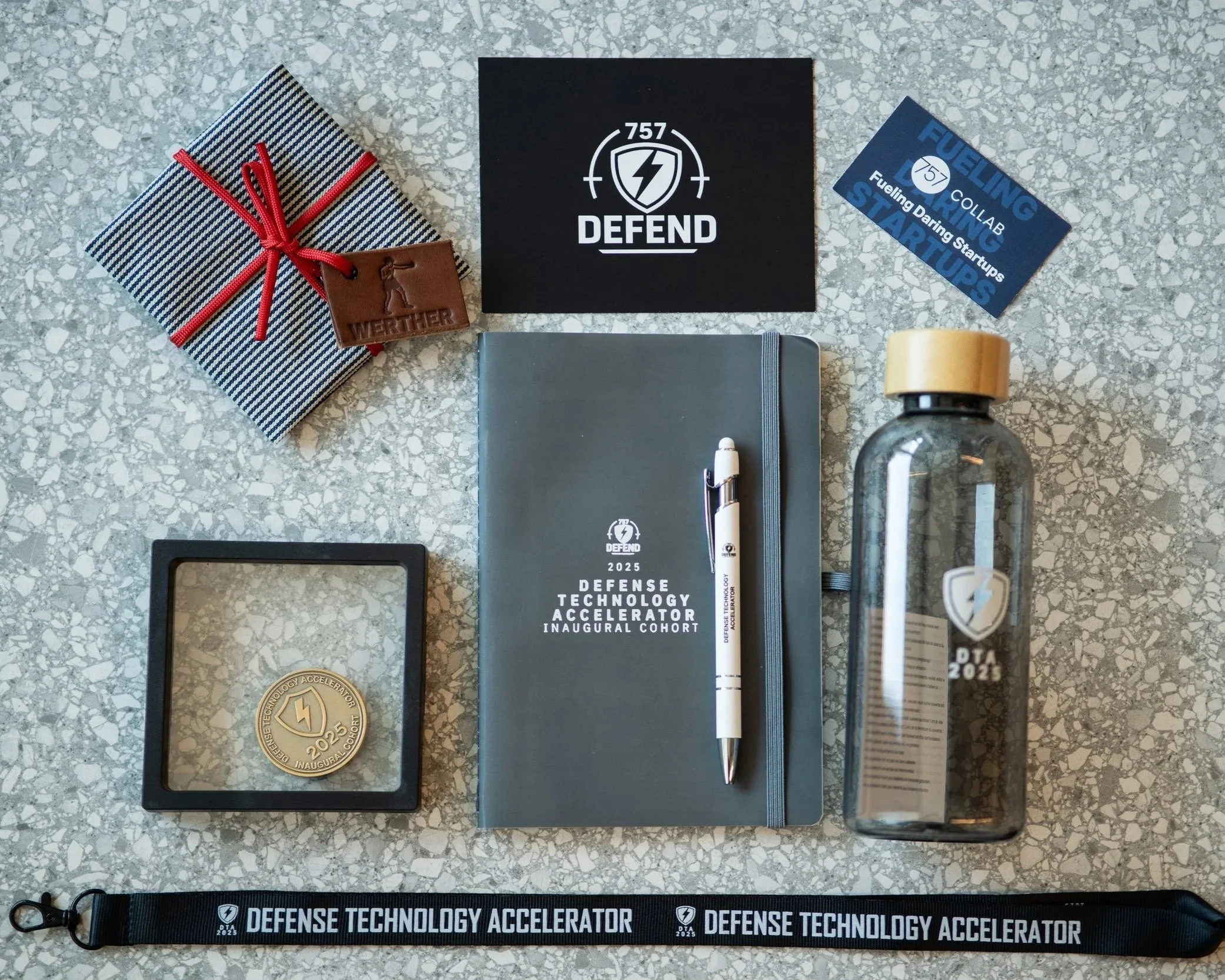 Collection of items including a wrapped gift, stickers, a notebook with a pen, a water bottle, a medal, a lanyard, and various branded materials on a speckled gray surface.