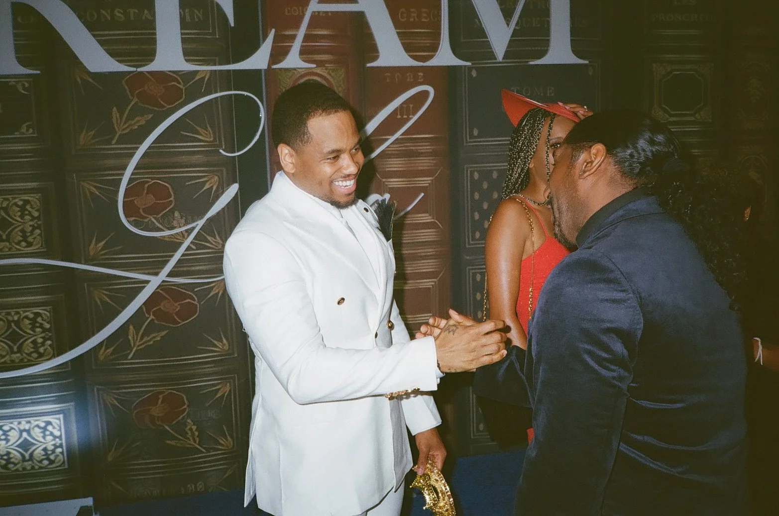 Two men greeting each other at a social event, one in a white suit and the other in a dark suit, with a woman in a red dress and hat in the background.