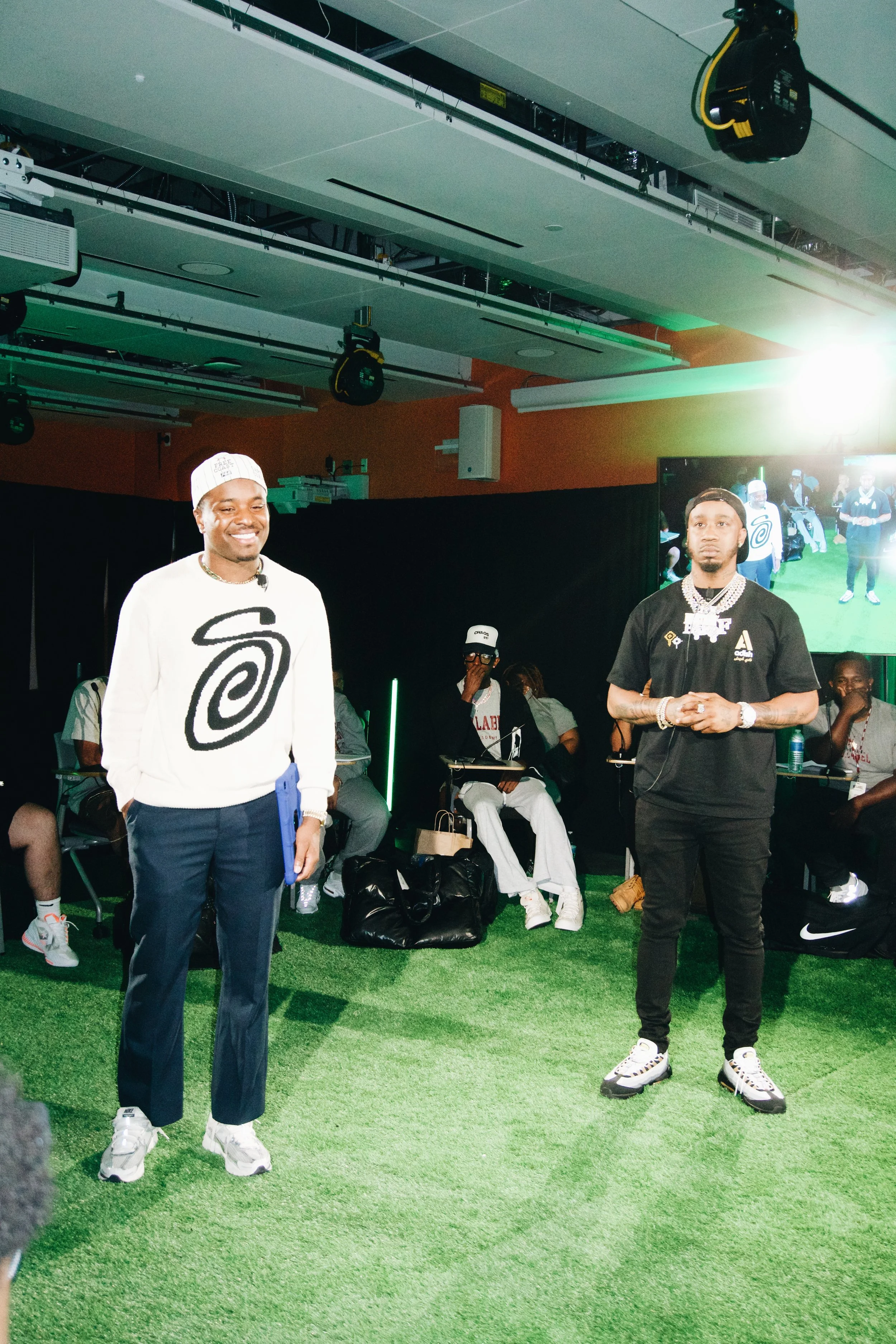 Two men stand in front of an audience at an indoor event, with a large screen behind them showing the same scene.