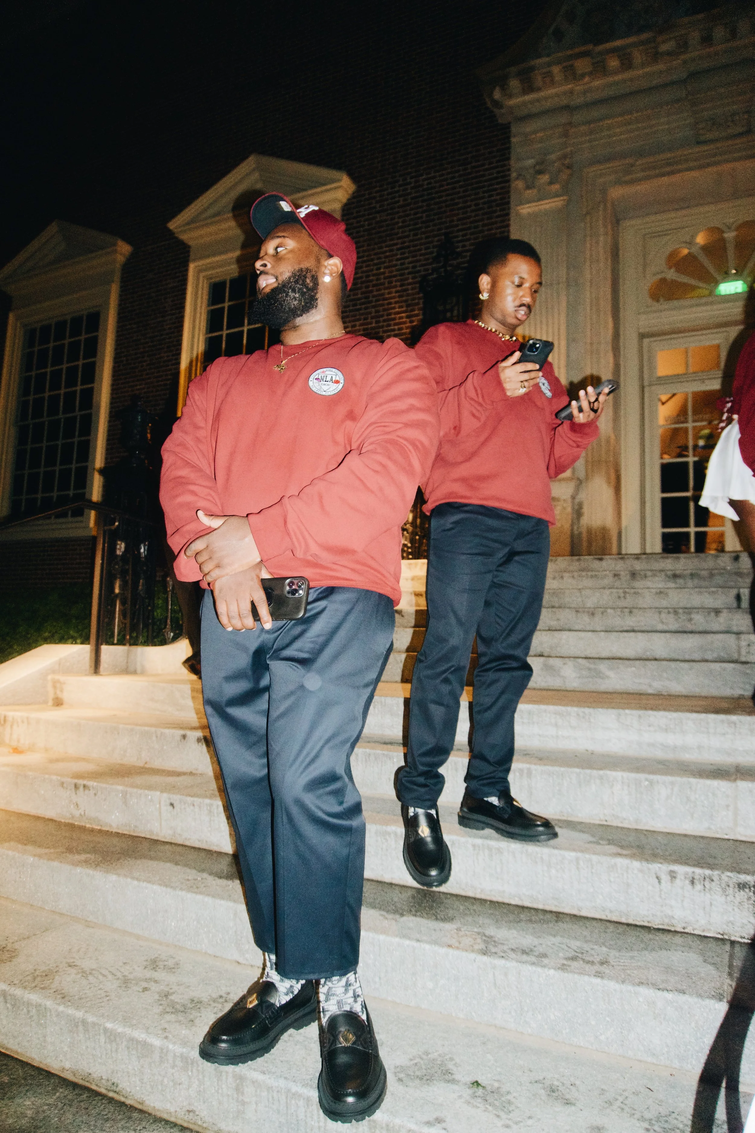 Two men wearing matching red jackets and dark pants standing on a staircase outside a building at night, both looking at their phones.