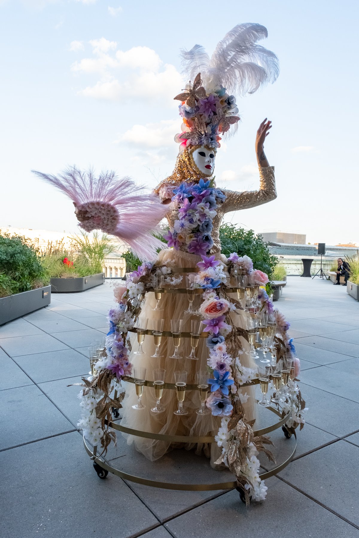 A person dressed in an elaborate costume with a mask, gold and floral decorations, feathers, and flowers, standing outdoors on a terrace with plants and a person sitting in the background.