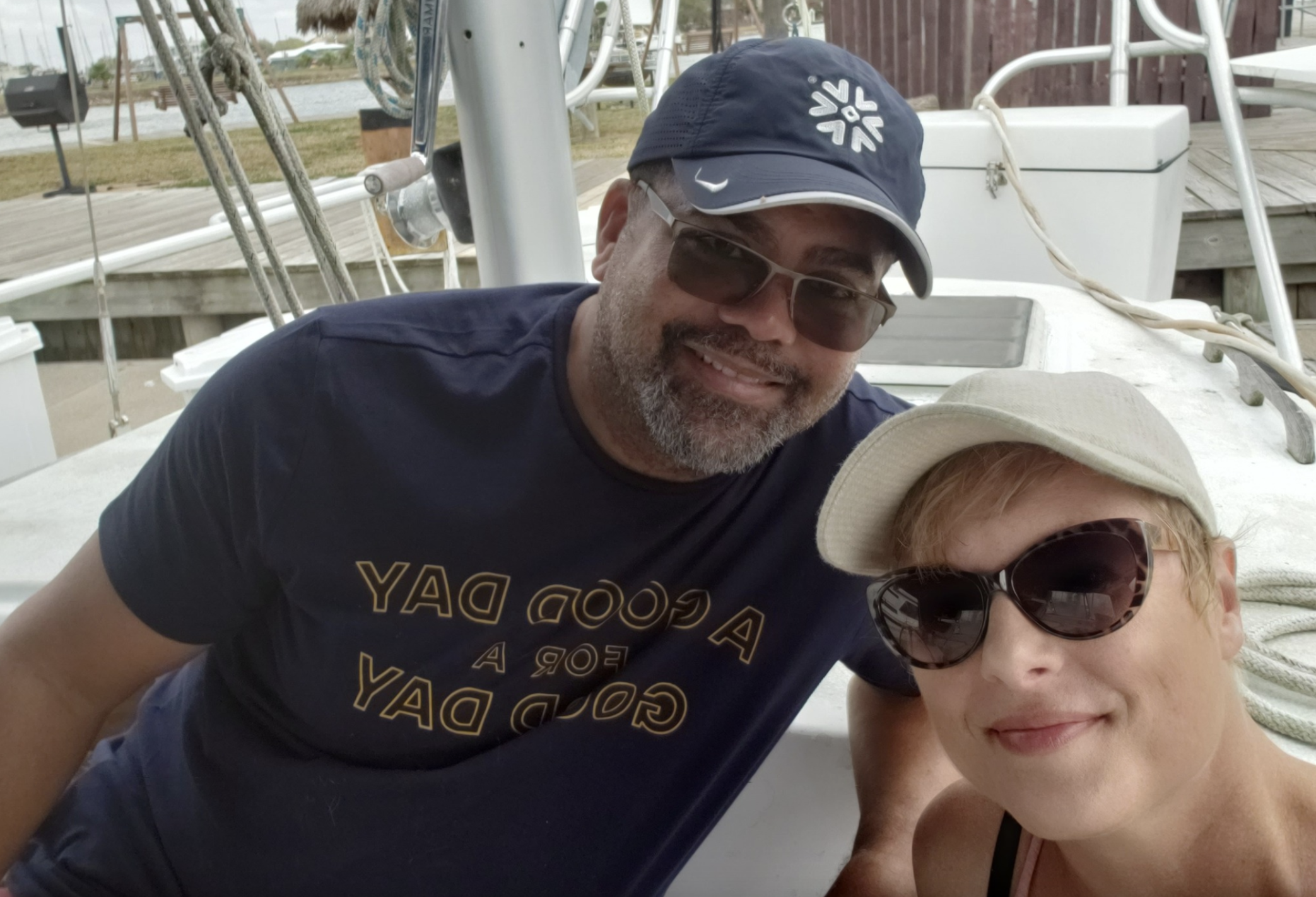 A man and woman taking a selfie on a boat, both wearing sunglasses and hats. The man has a beard, is smiling, and wearing a navy blue cap and T-shirt. The woman is smiling, wearing a beige cap and large sunglasses.