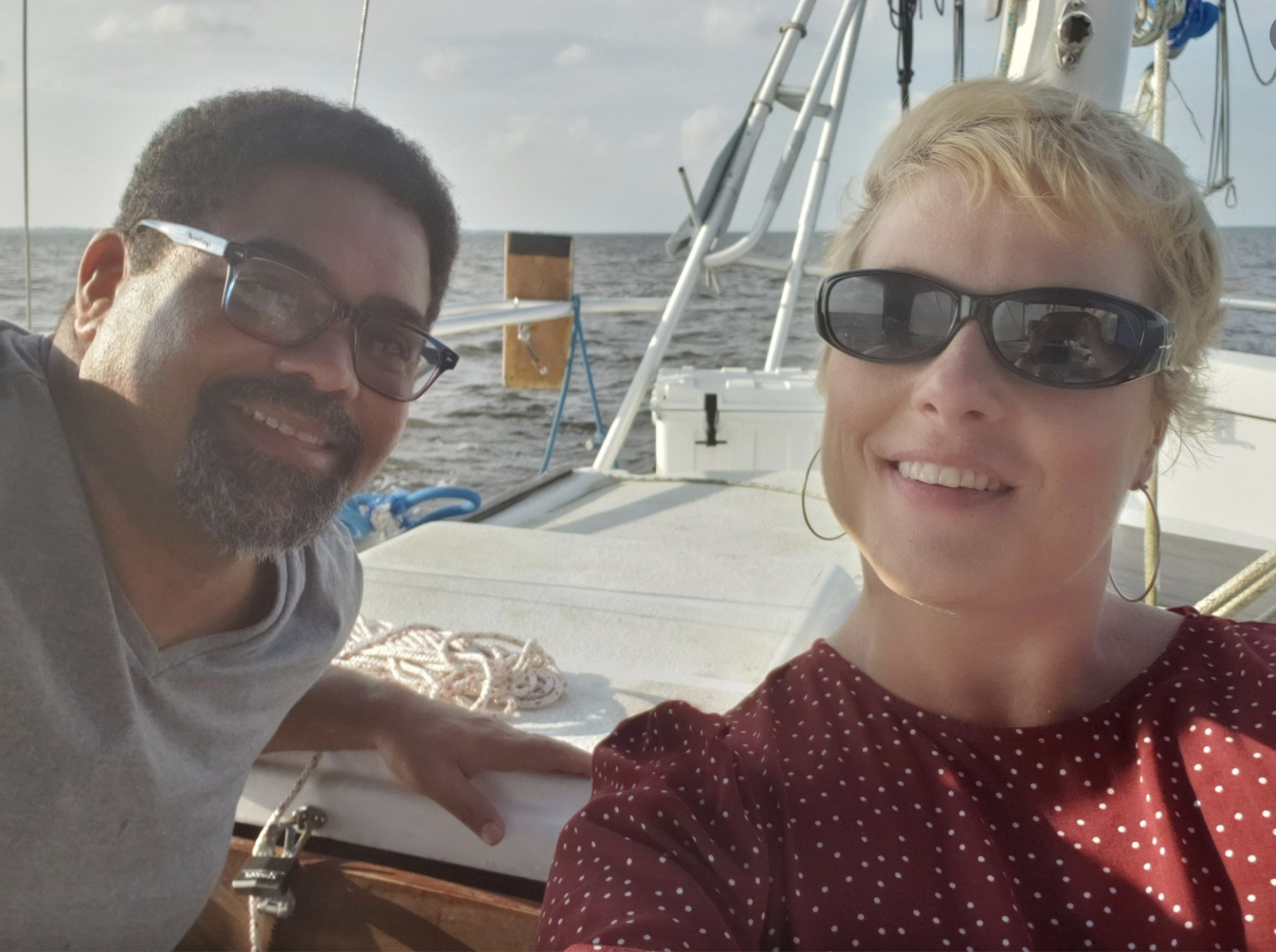 A smiling man with glasses and a beard and a smiling woman with sunglasses and hoop earrings on a boat at sea.