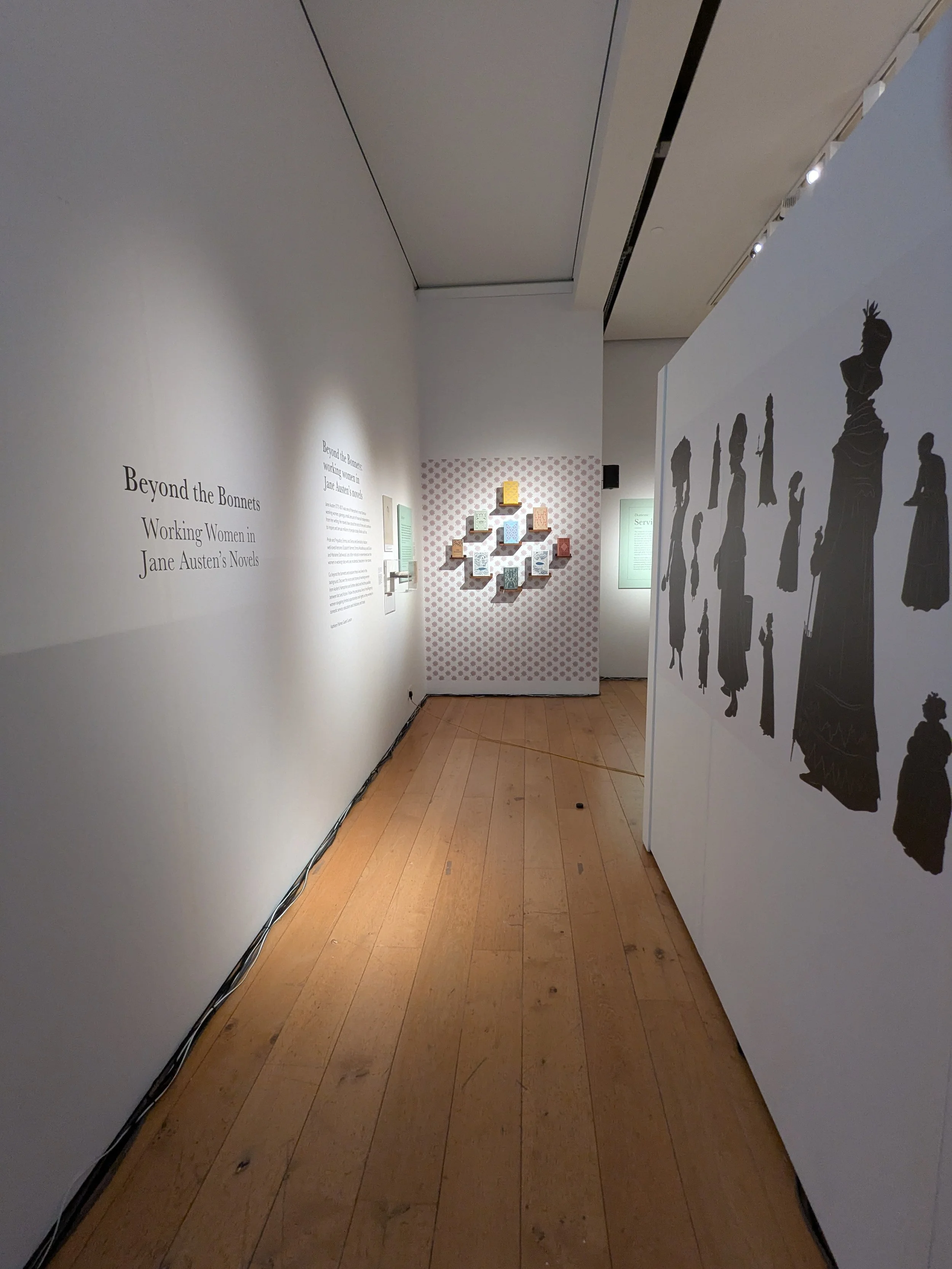 Exhibition entrance. Introductory text on the left wall. Ahead the far wall has a display of Austen's novels. The right wall has silhouettes of Regency working women.