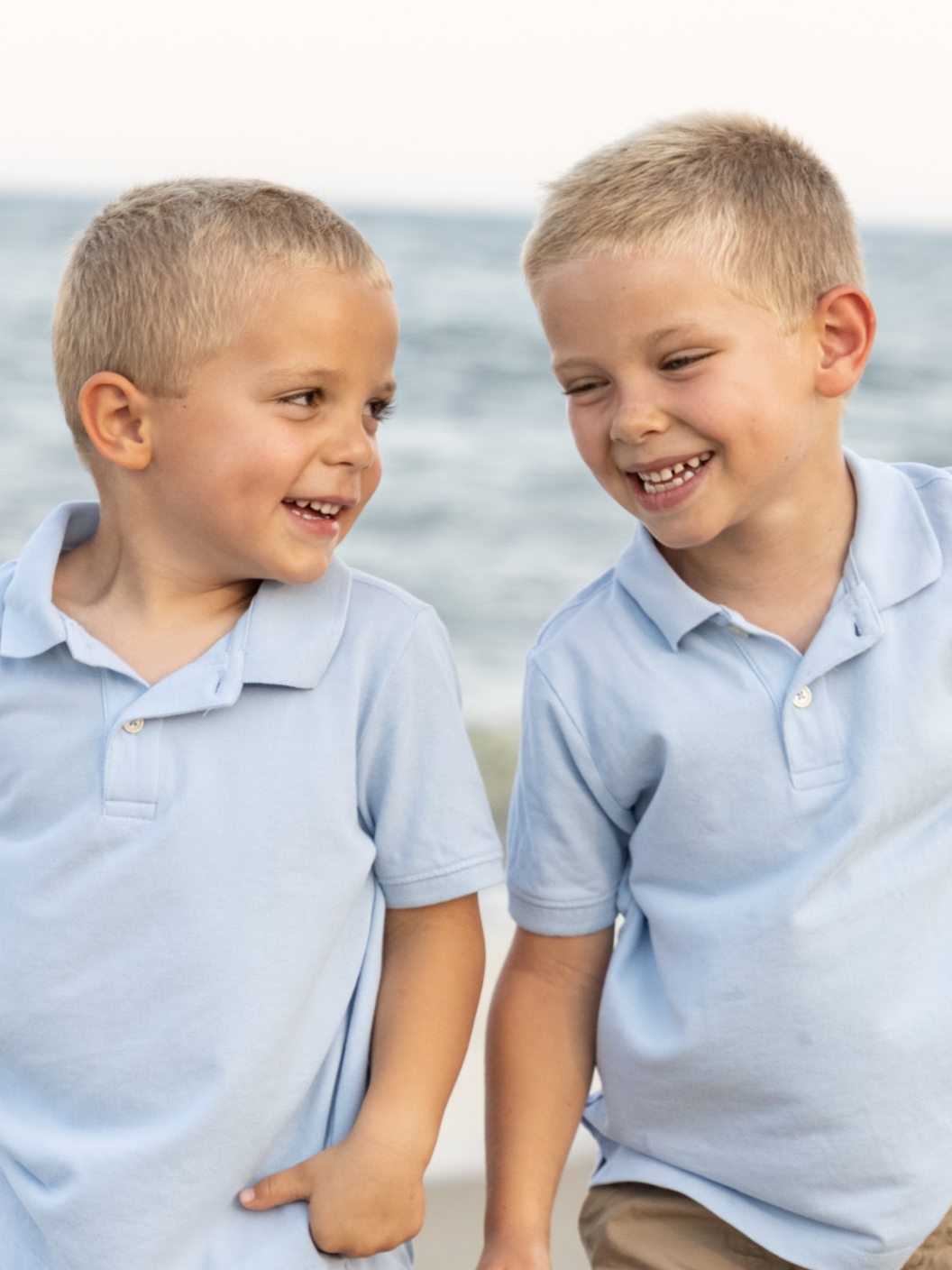 Two young boys wearing light blue polo shirts and khaki shorts are playing and smiling on the beach near the ocean.