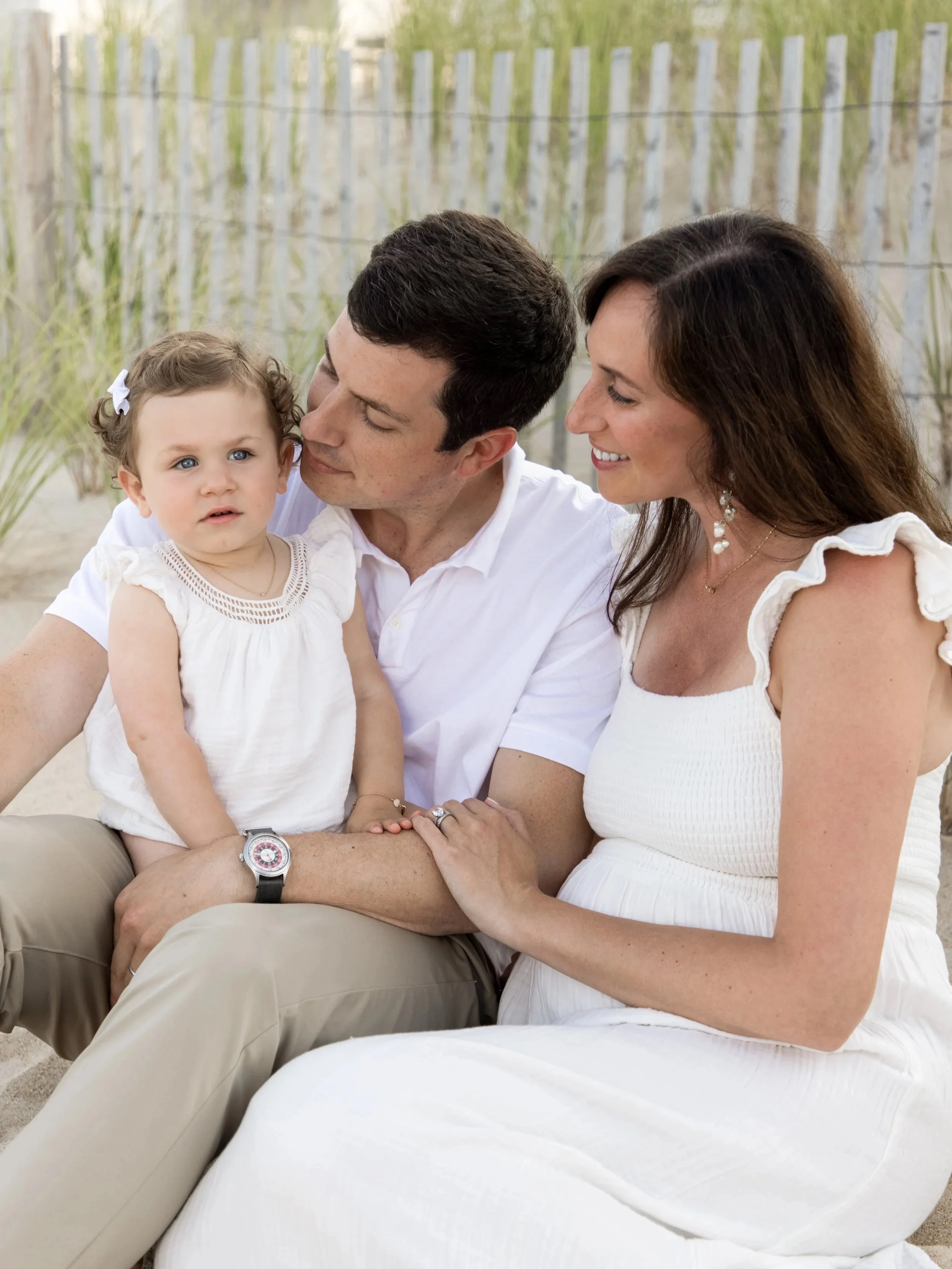 Family of three sitting outdoors on the sand, the father holding a young girl with blonde hair, and the mother sitting close to them, all smiling and dressed in white.
