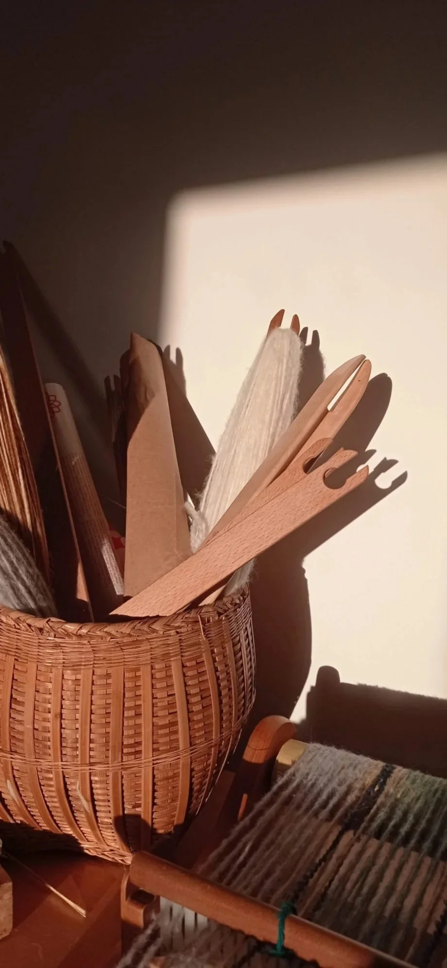 A wicker basket holding wooden stir sticks and paper napkins, with sunlight casting shadows on a beige wall.