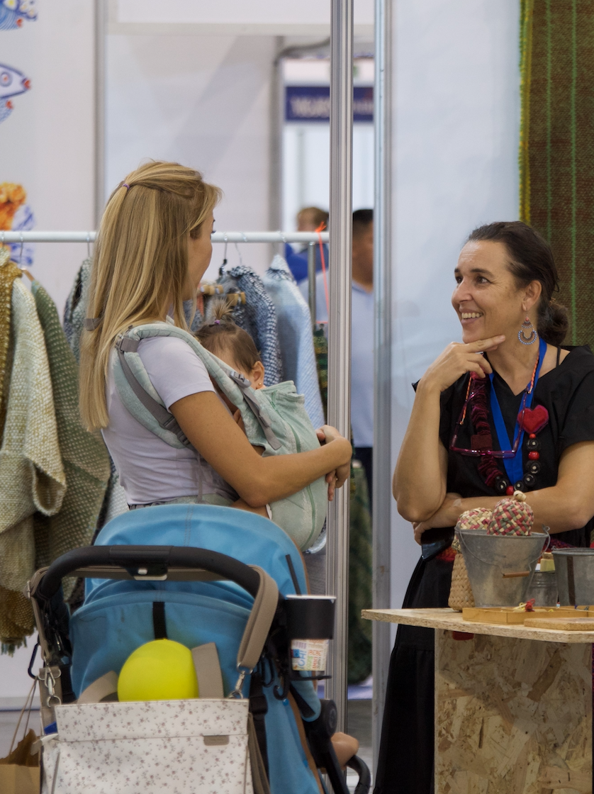 A man sitting on a beige cushioned bench against a beige wall, wearing a blue short-sleeve sweater and beige pants, looking to his left.